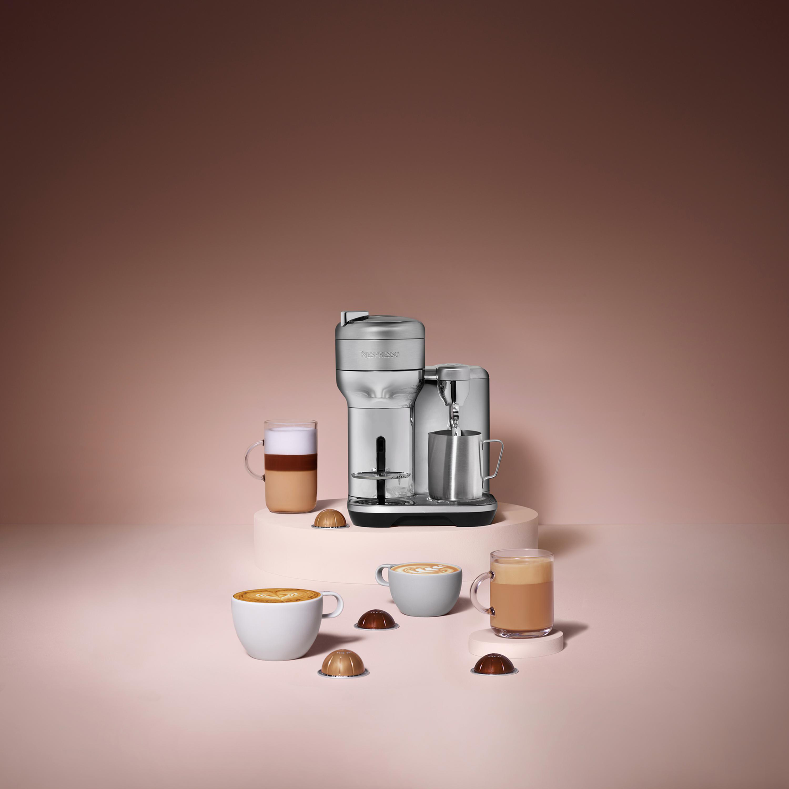 Eine Kaffeemaschine, Tassen mit Kaffee und Kapseln auf einer rosa Oberfläche.