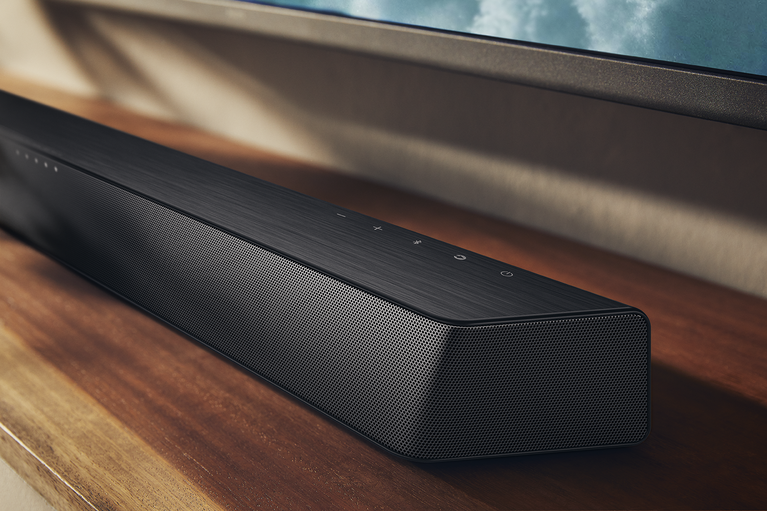 Een zwarte soundbar staat op een houten oppervlak. Het heeft een geperforeerde voorkant en bedieningsknoppen. Een tv staat op de achtergrond.