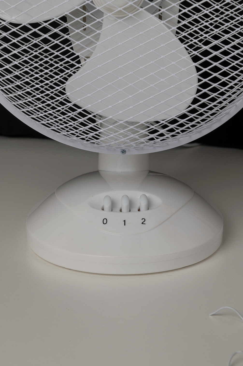 Ventilateur blanc avec réglages de vitesse 0, 1 et 2. Fond noir, table blanche.