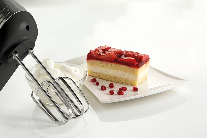 Egy fekete kézi mixer egy szelet tortával, tejszínhabbal és gránátalmamaggal.