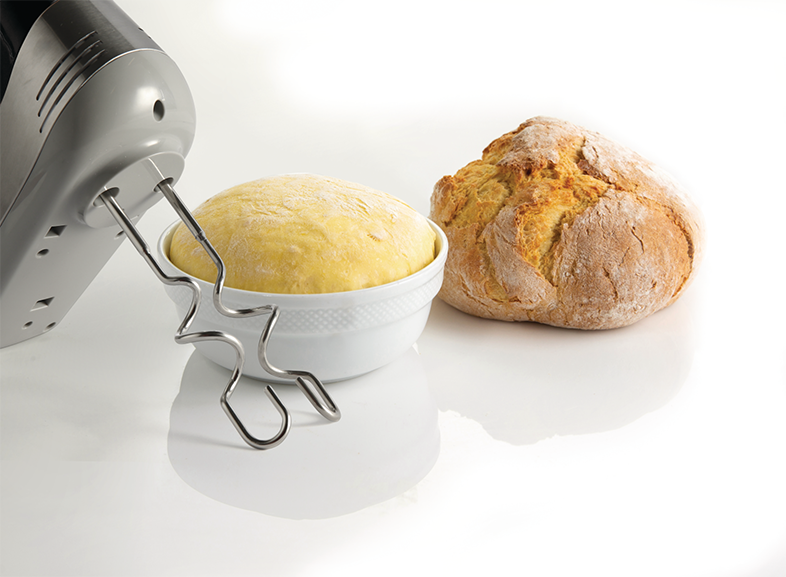 Kézi mixer, amely tésztát kever egy tálban egy kenyér mellett, fehér háttéren.