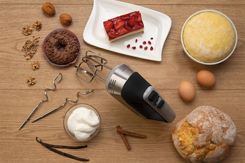 Kézi mixer hozzávalókkal: sütemény, tészta, krém, tojás, dió és fűszerek a fa felületen.