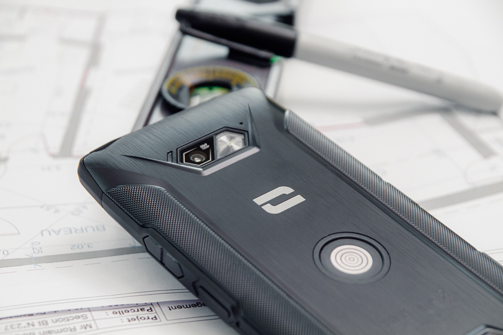 Un téléphone noir robuste sur un plan, avec un stylo et un niveau.