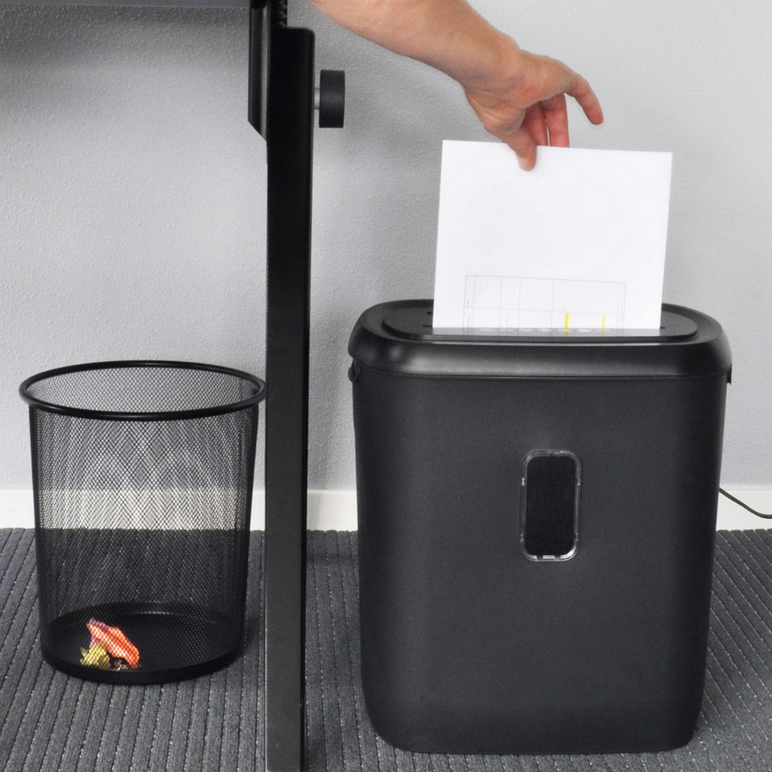 A person putting a paper into a black paper shredder next to a trash can.