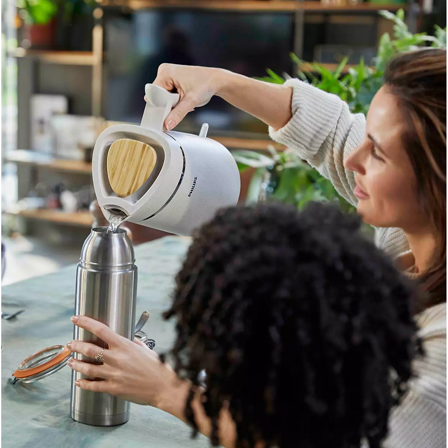A woman pouring hot water from a kettle into a steel thermos. Another woman is watching.