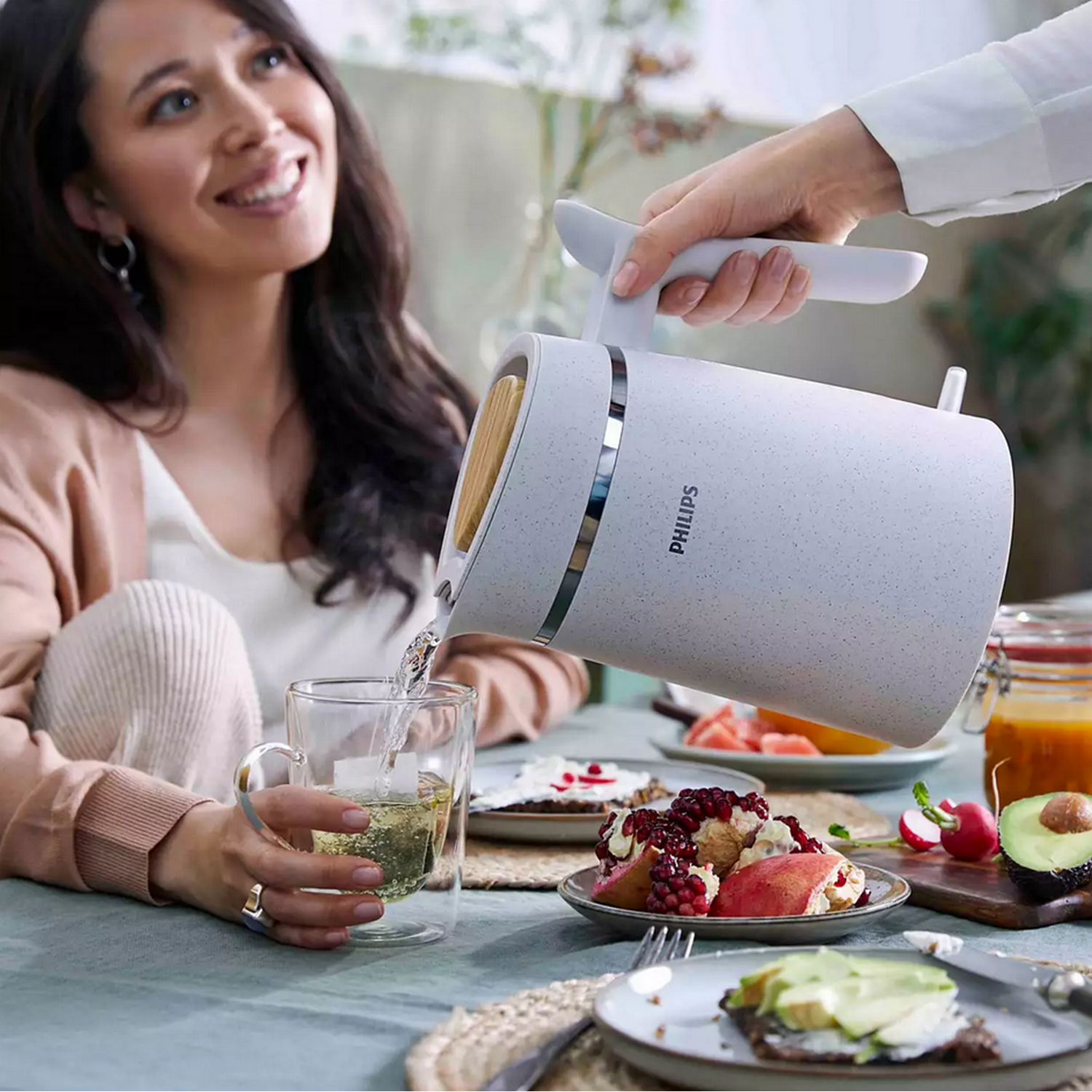 Woman pouring water from a white kettle into a glass, table setting.