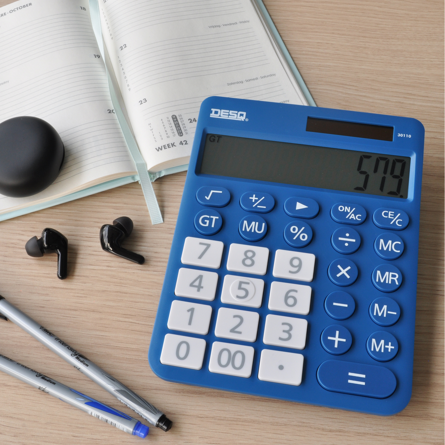 A desk with a calculator, headphones, pens, and a planner. A person is writing on a paper.