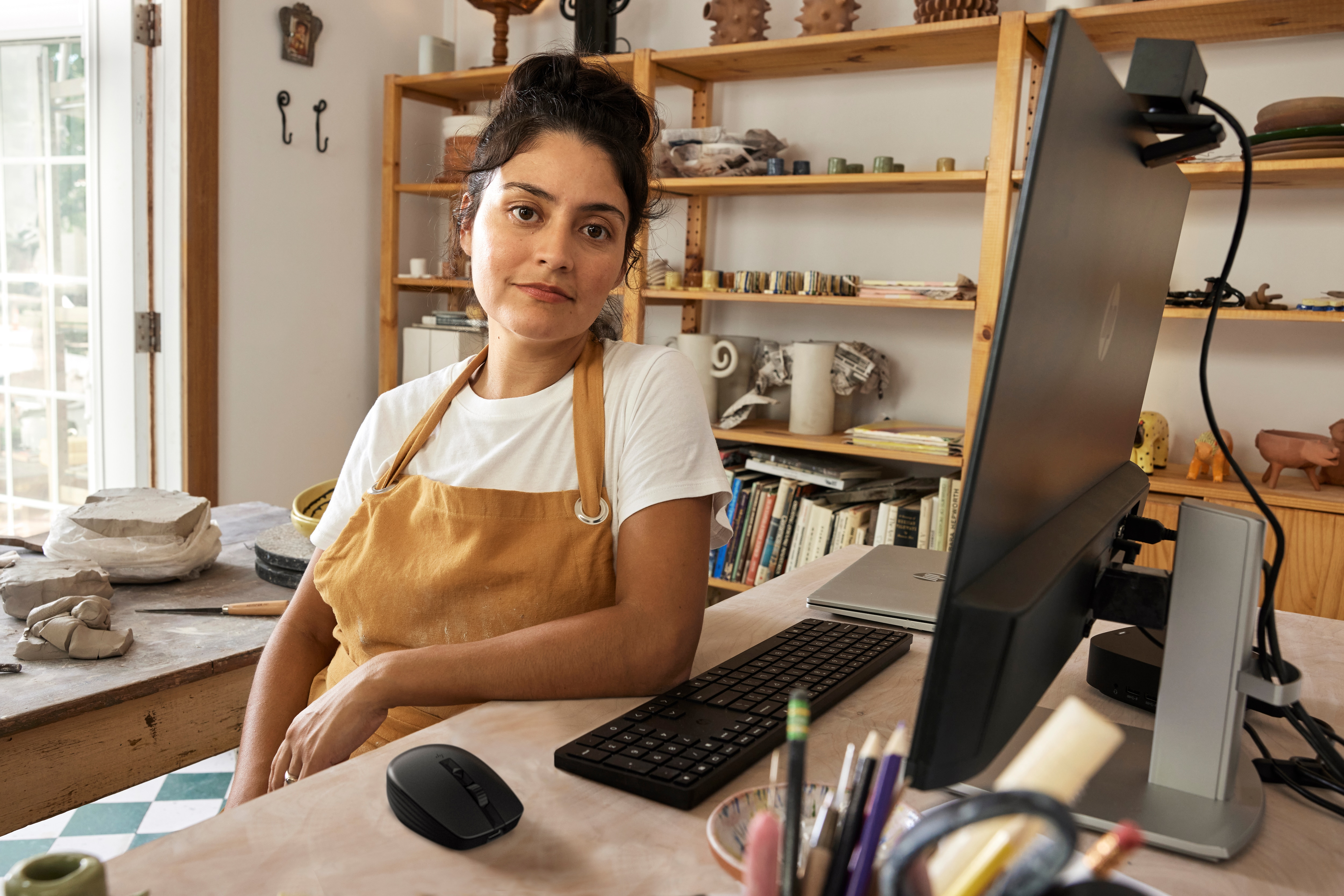 Eine Frau sitzt an einem Schreibtisch in einem Atelier. Ein Computermonitor, Tastatur, Maus und Kunstutensilien sind vorhanden.