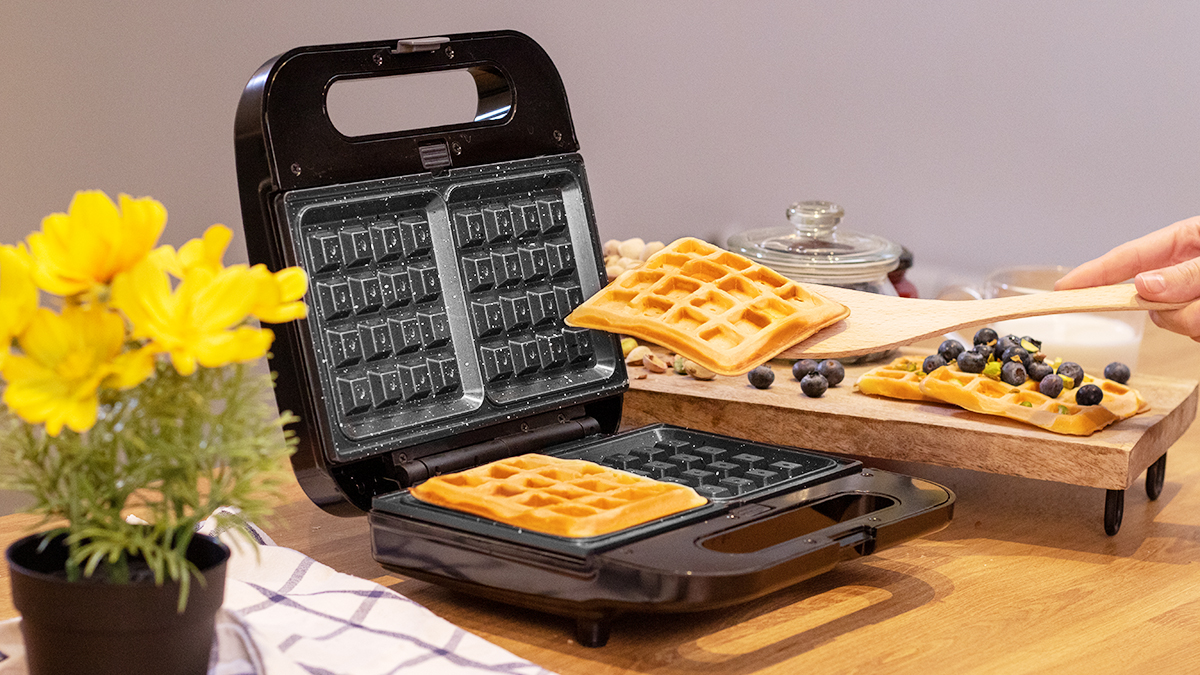 Una máquina para hacer gofres con dos gofres, uno que se quita y otro listo, con una tabla de madera.