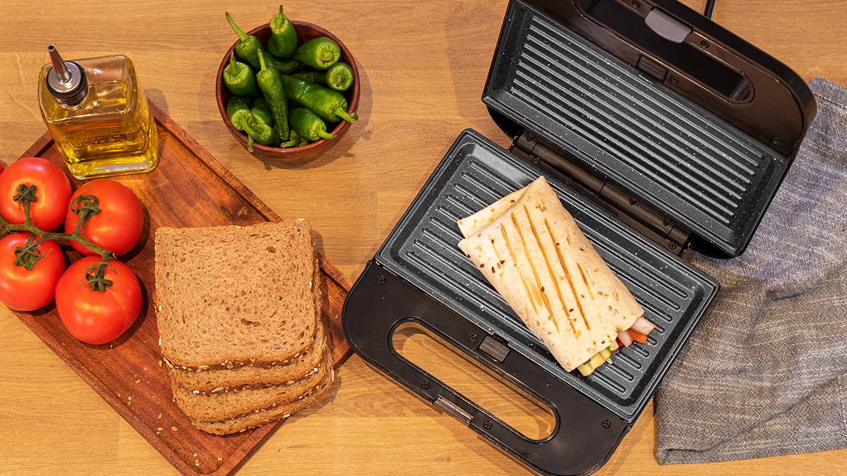 Sandwichera con un envoltorio dentro, con tomates, pan y pimientos sobre una superficie de madera.