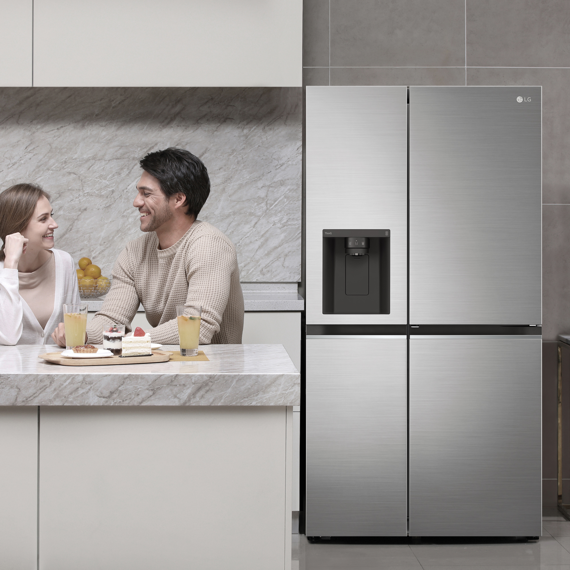 Una pareja está sentada en una encimera de la cocina con pastel, jugo y un refrigerador LG.