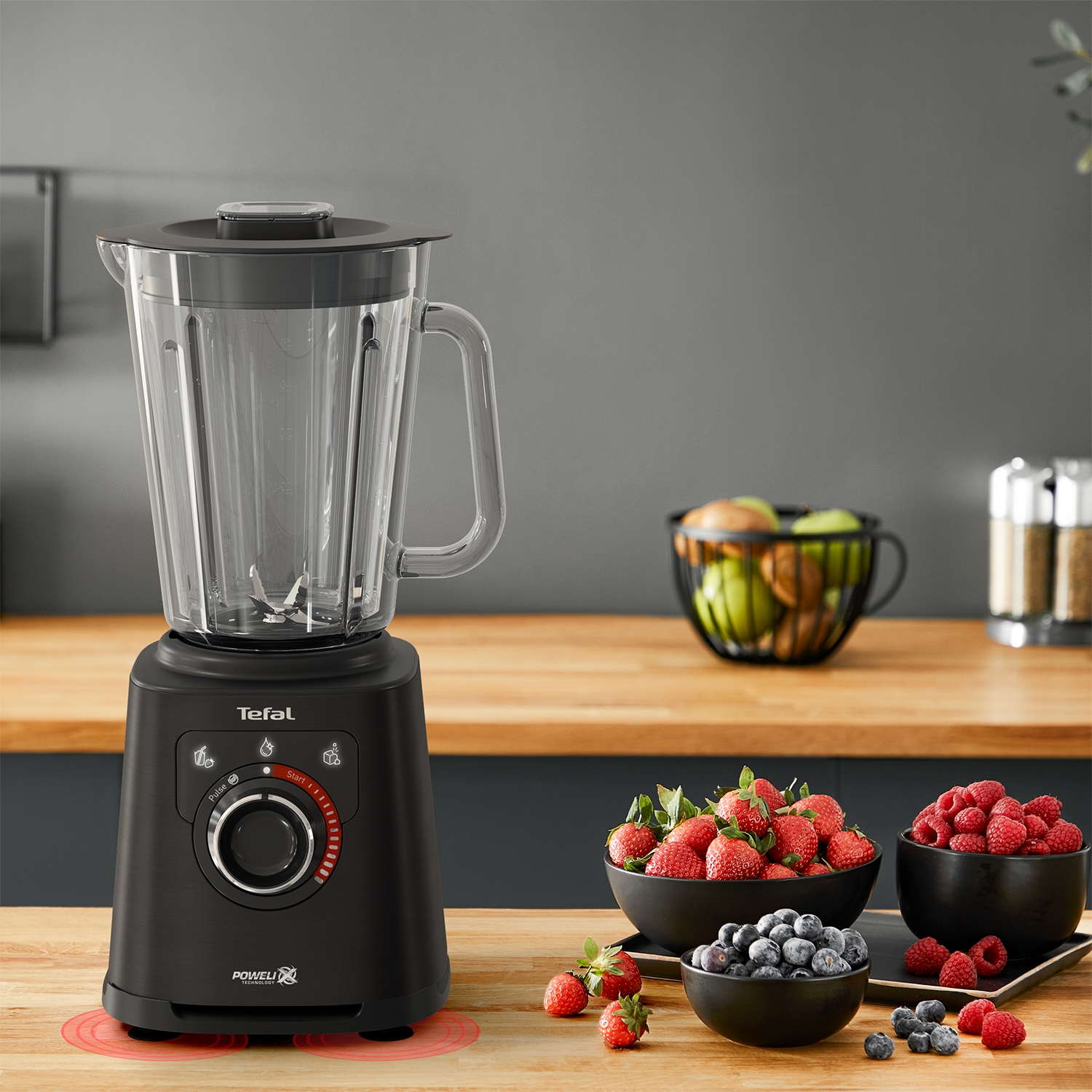Black blender with fruit bowls. Strawberries, blueberries, and raspberries are on a wooden table.