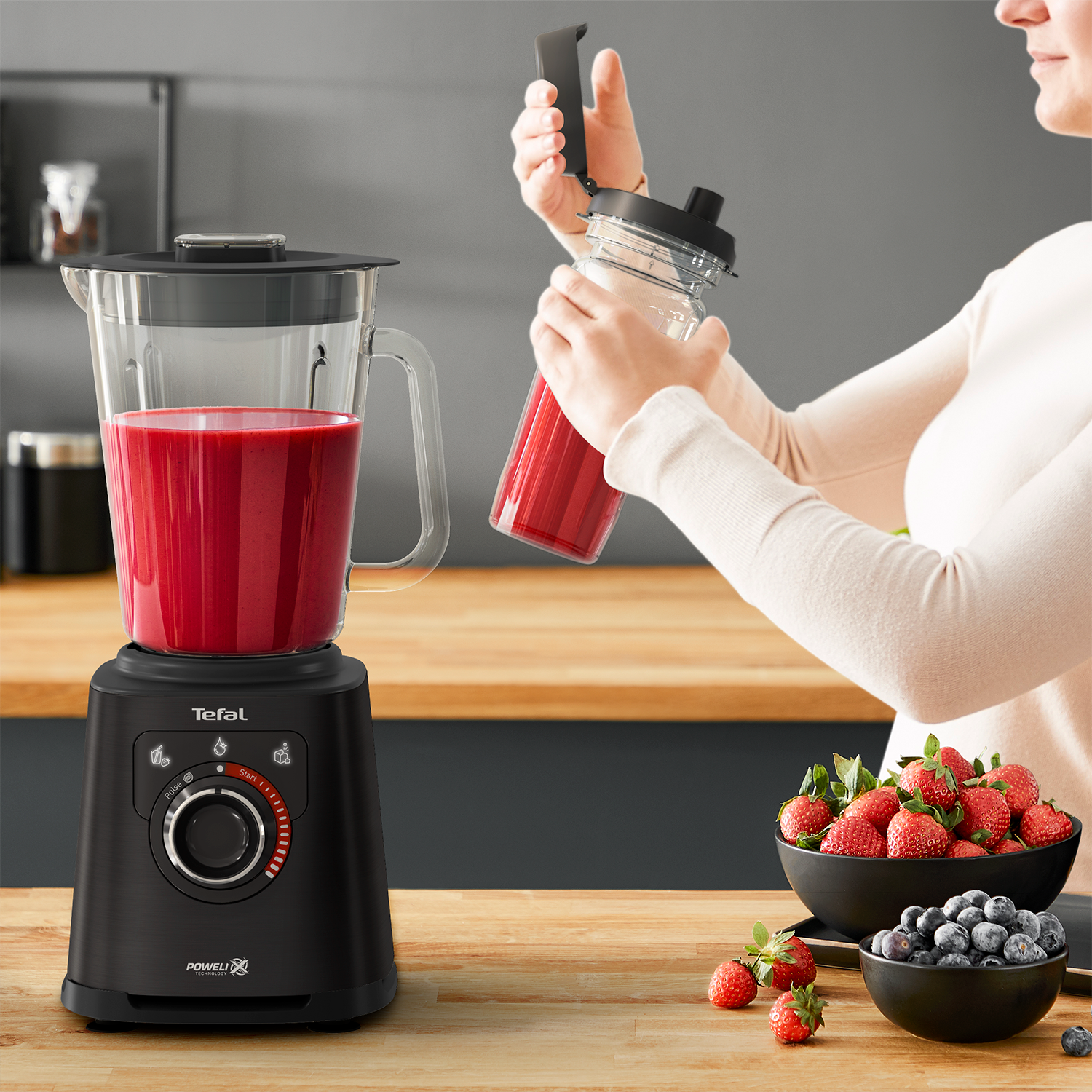 Black blender with red smoothie, woman holding a jar. Strawberries and blueberries on wooden table.