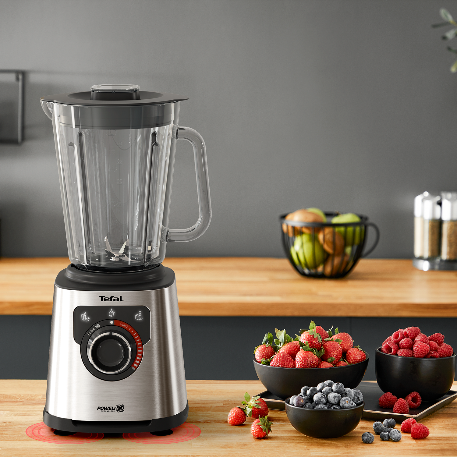Silver blender on a wooden table. Bowls of berries in the foreground, and a black kitchen.