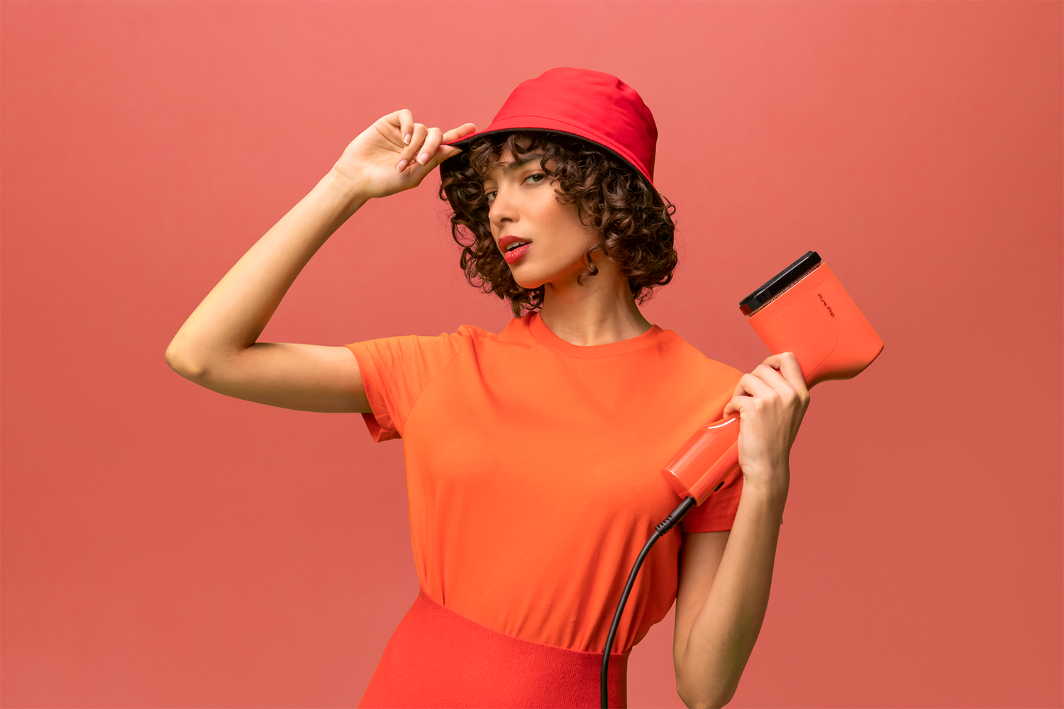 Woman in orange outfit holding a hair dryer and a red hat.