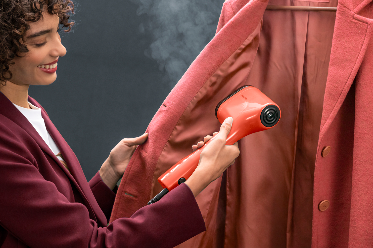 Woman steams a pink coat with an orange garment steamer. She is smiling.