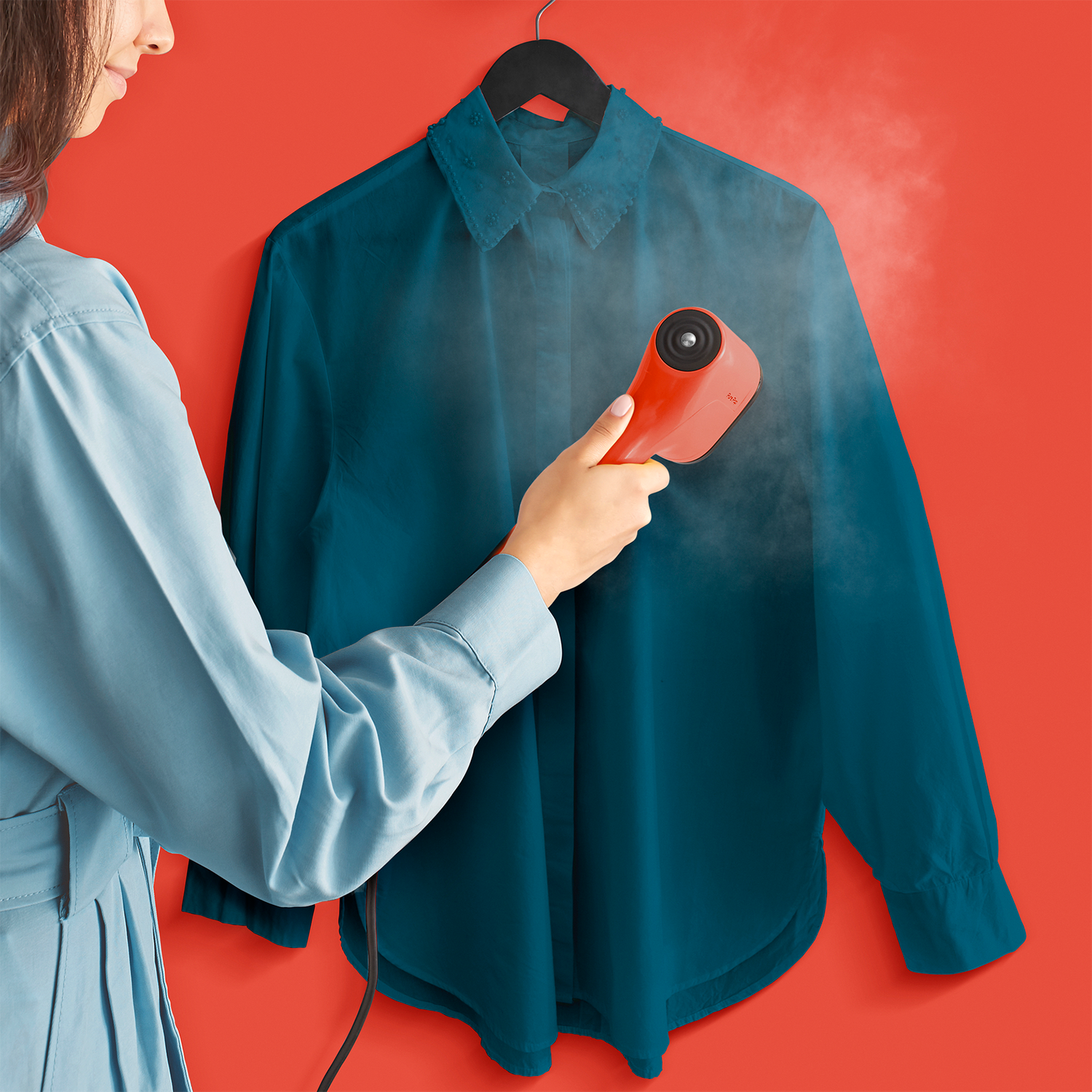 Woman ironing a teal shirt on a hanger with a red steamer, on a red background.