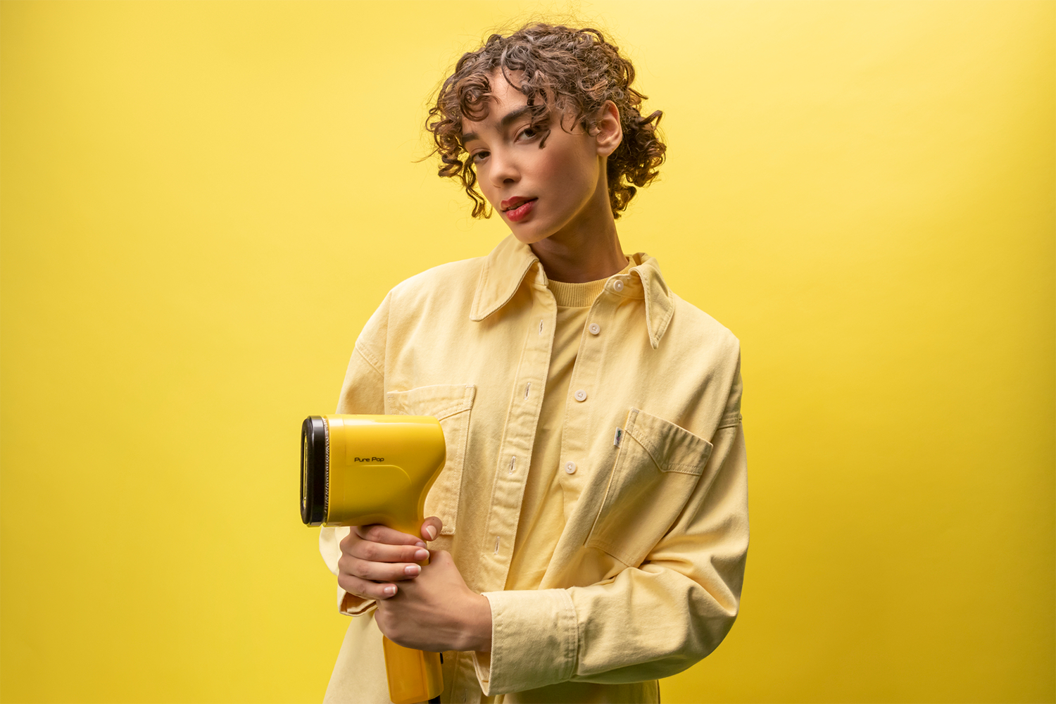 Young woman with curly hair holding a yellow hair dryer on a yellow background.