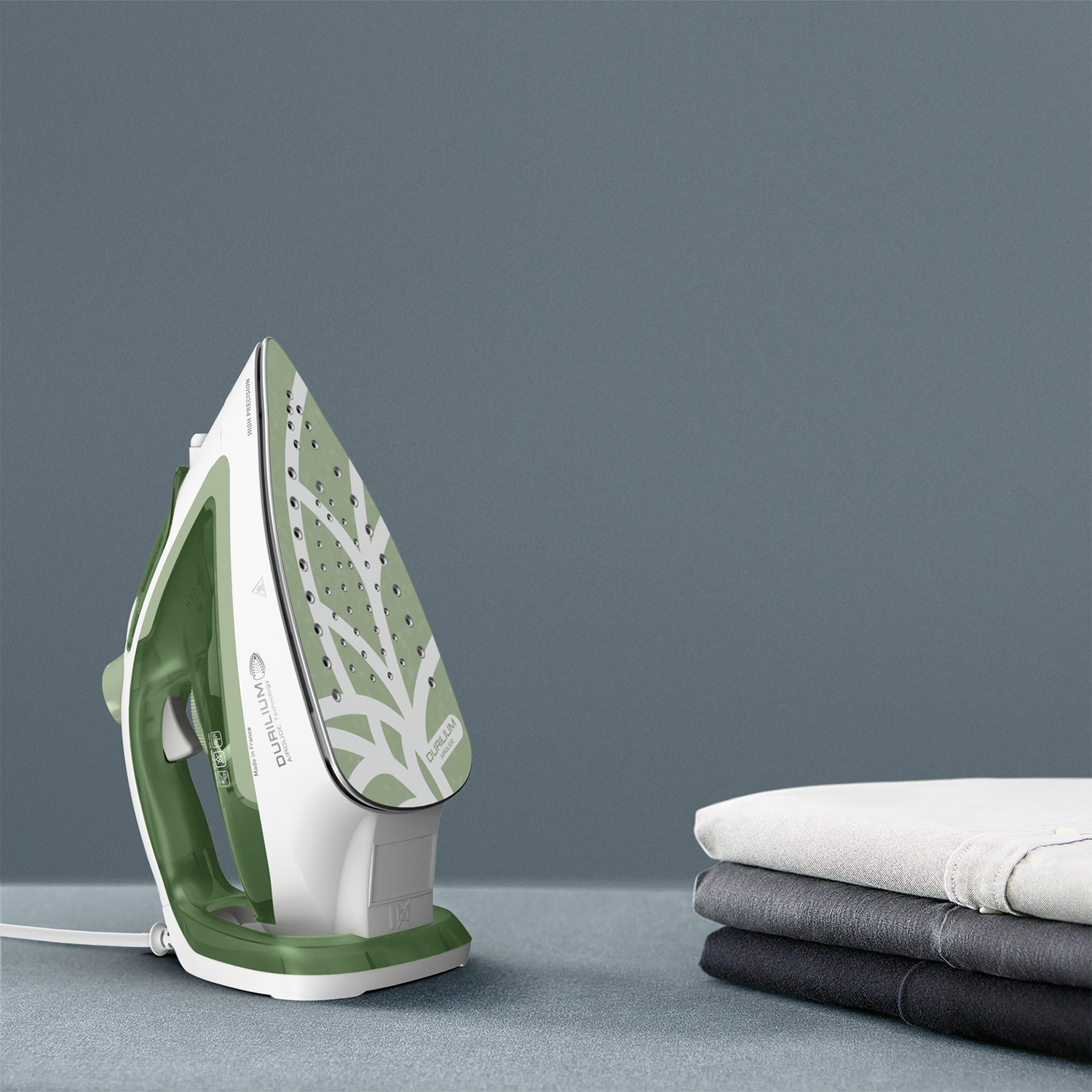 A green and white iron with clothes stacked on a table.