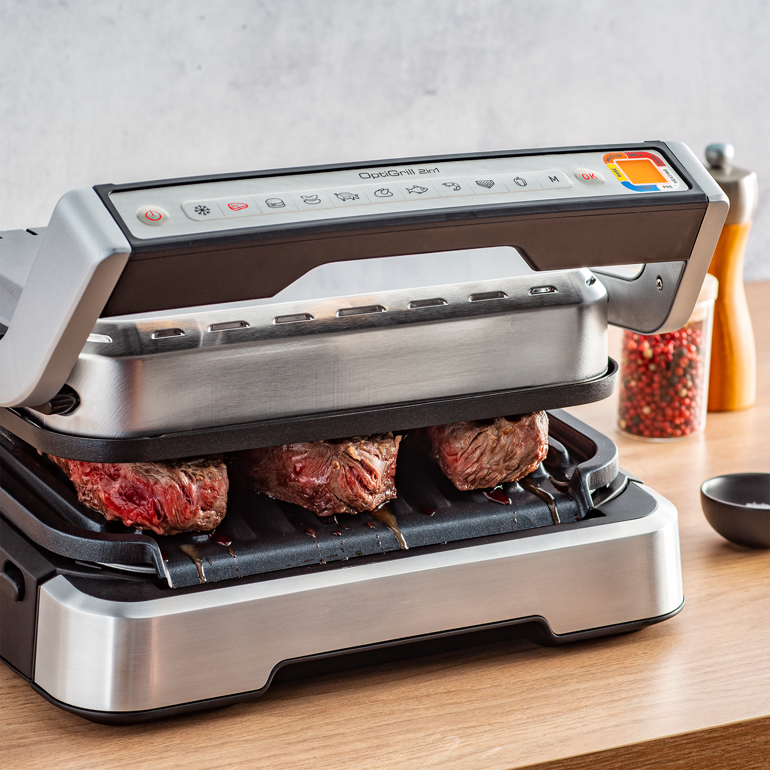 A silver and black OptiGrill with three steaks cooking. The grill is on a wooden surface with spices.