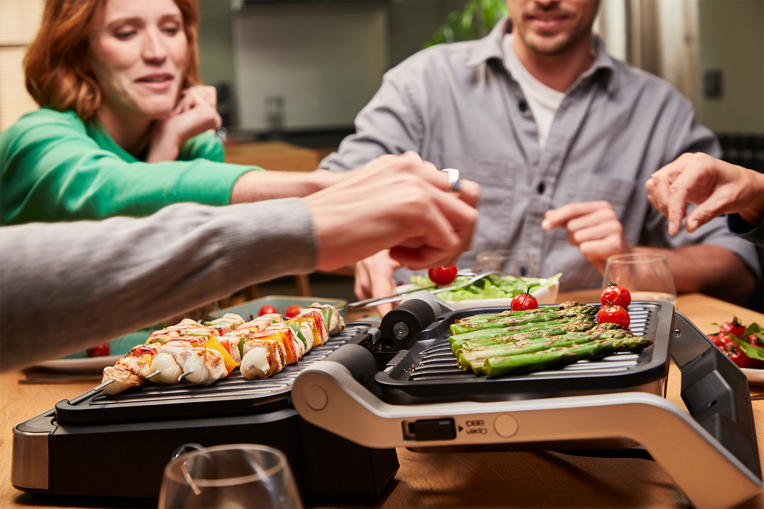 A group of people around a table grilling food on an electric grill, with kebabs and asparagus.