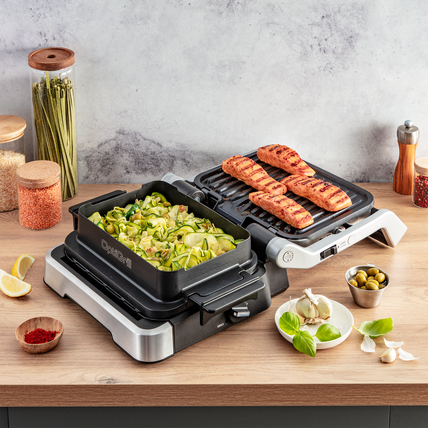 Twee elektrische grills met eten: gegrilde zalm en courgette. Ingrediënten zoals olijven en knoflook in de buurt.