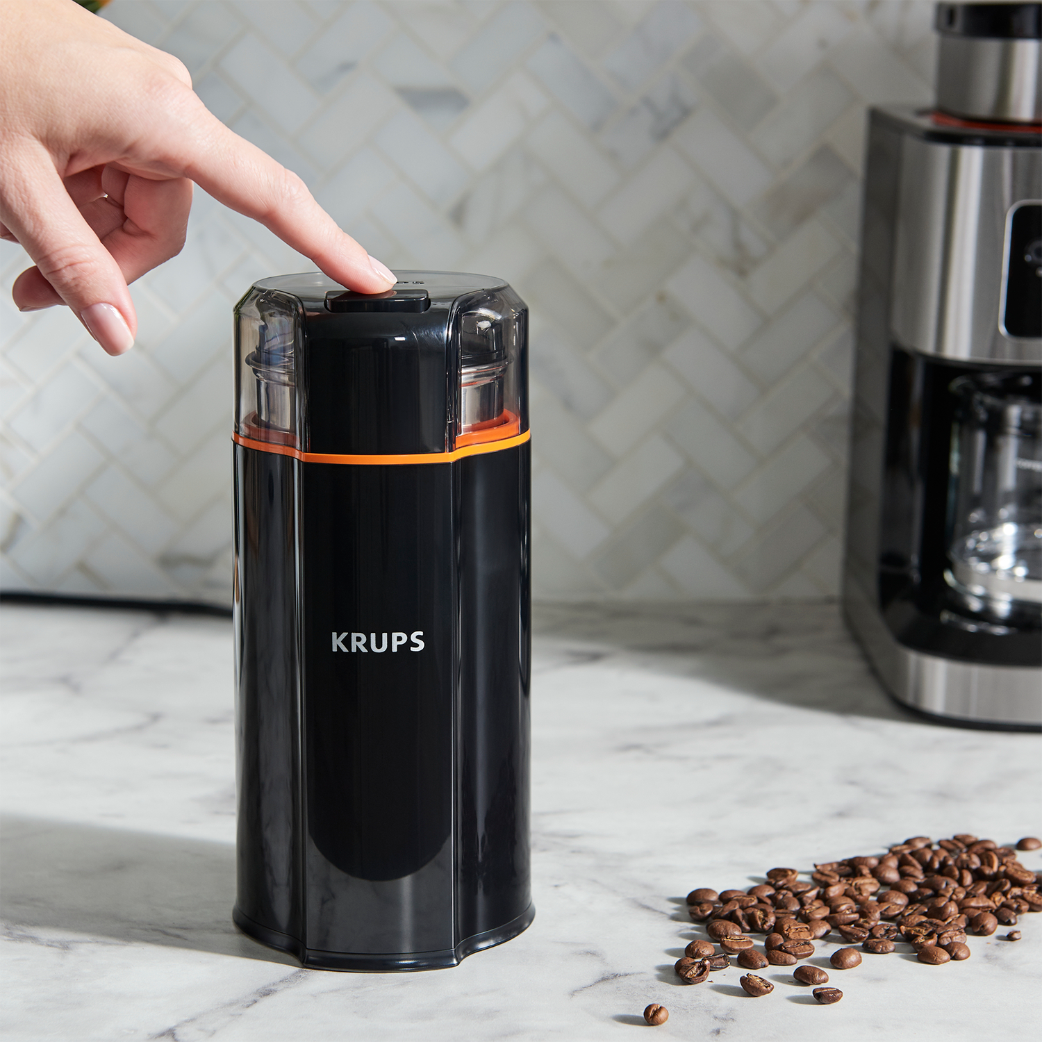 Person presses a button on a black Krups coffee grinder. Coffee beans are scattered nearby, on a marble surface.