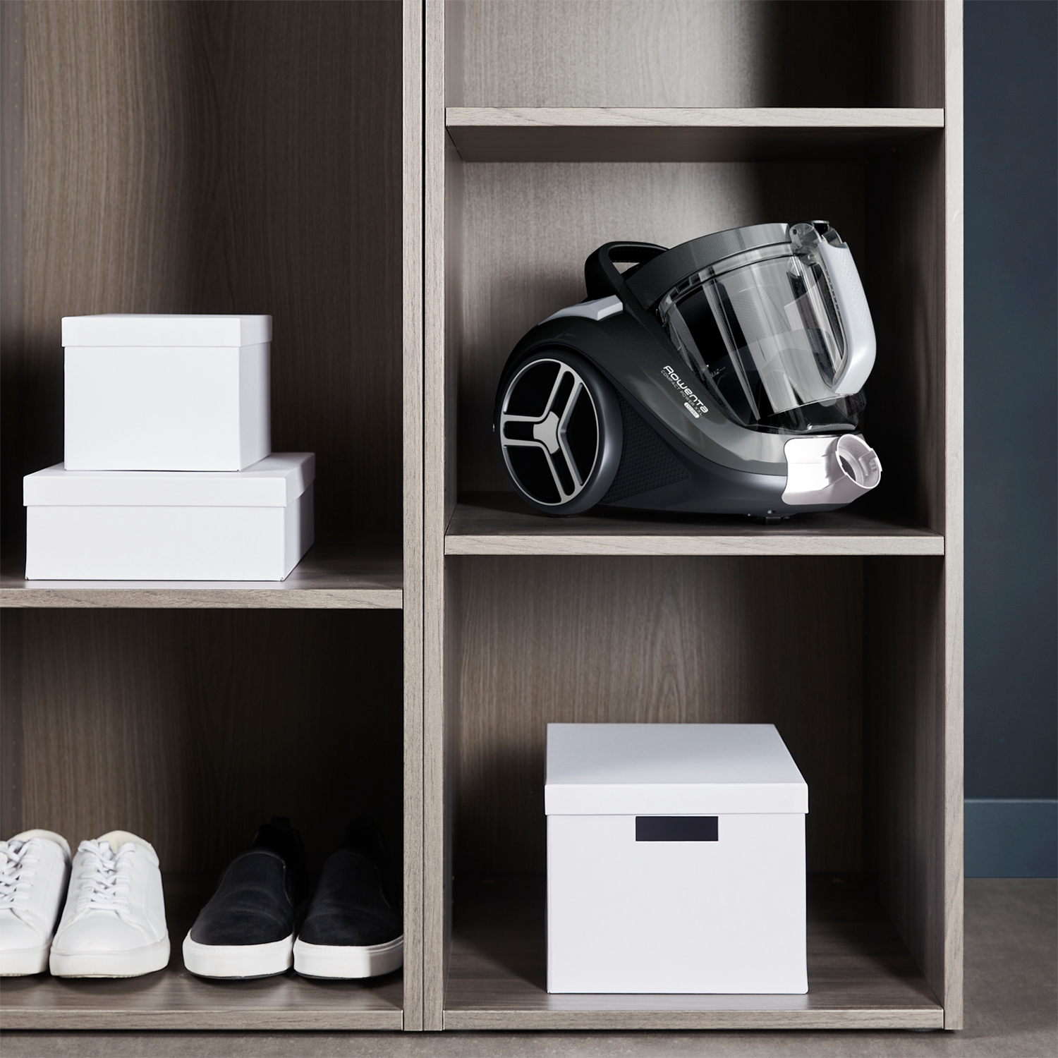 A vacuum cleaner sits on a shelf in a wooden cabinet with white boxes and shoes nearby.