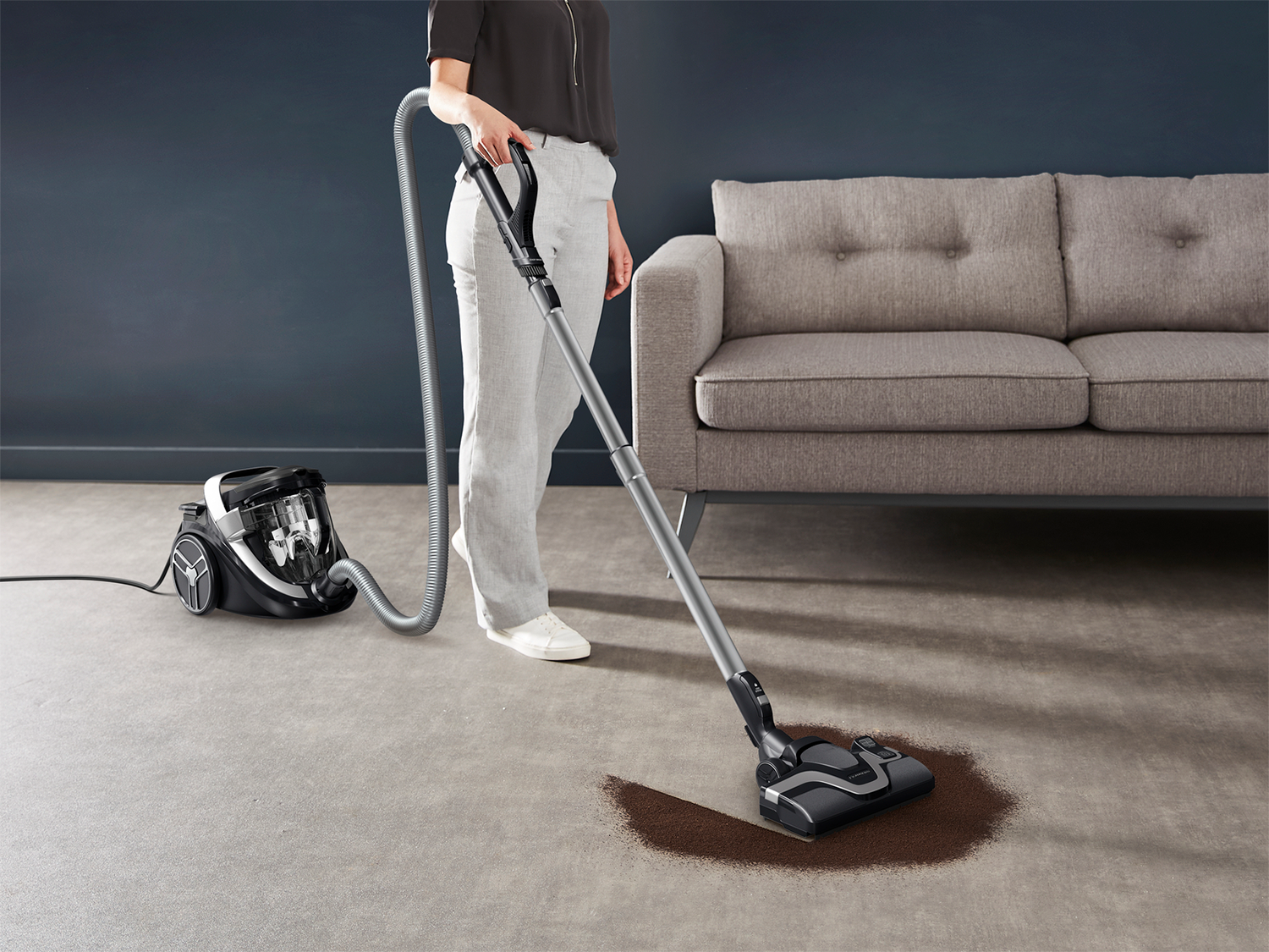 A woman vacuums a brown stain on a gray carpet in a room. A gray couch is behind her.