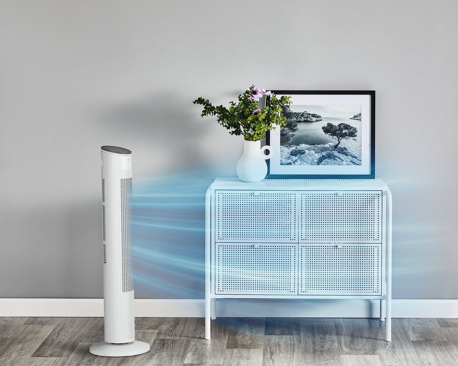 Un ventilateur blanc souffle de l'air vers une armoire blanche avec une image et un vase. Sol en bois et mur gris en arrière-plan.