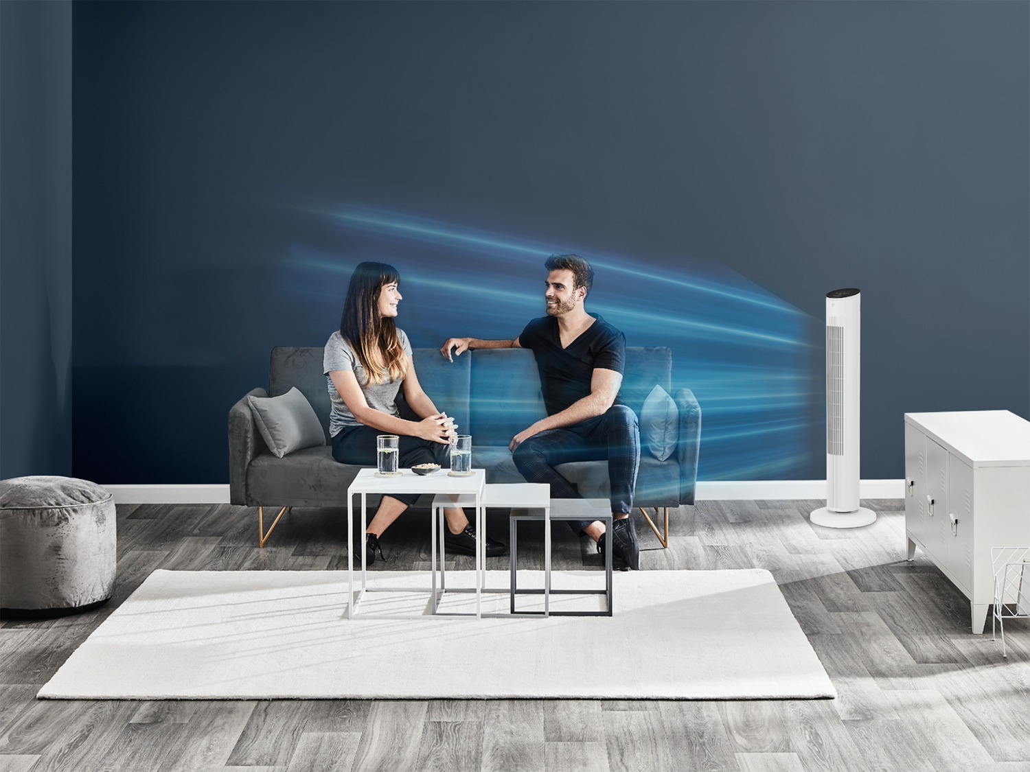 Un homme et une femme sont assis sur un canapé, en train de parler. Un ventilateur blanc est à droite. Une table contient des verres d'eau. Un tapis blanc est au sol.