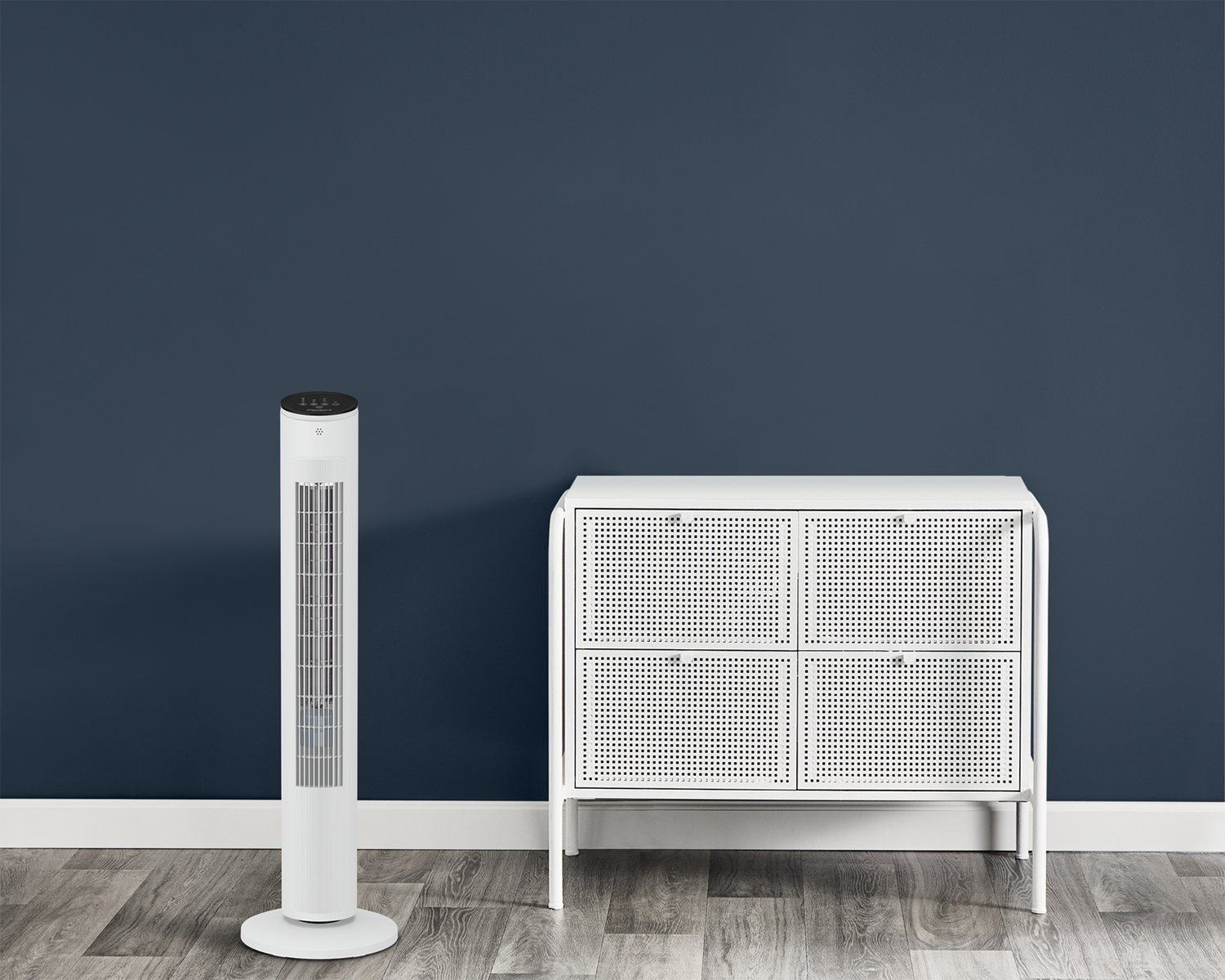 Un ventilateur tour blanc et un meuble blanc contre un mur bleu foncé sur un sol en bois. Les deux sont des designs modernes.