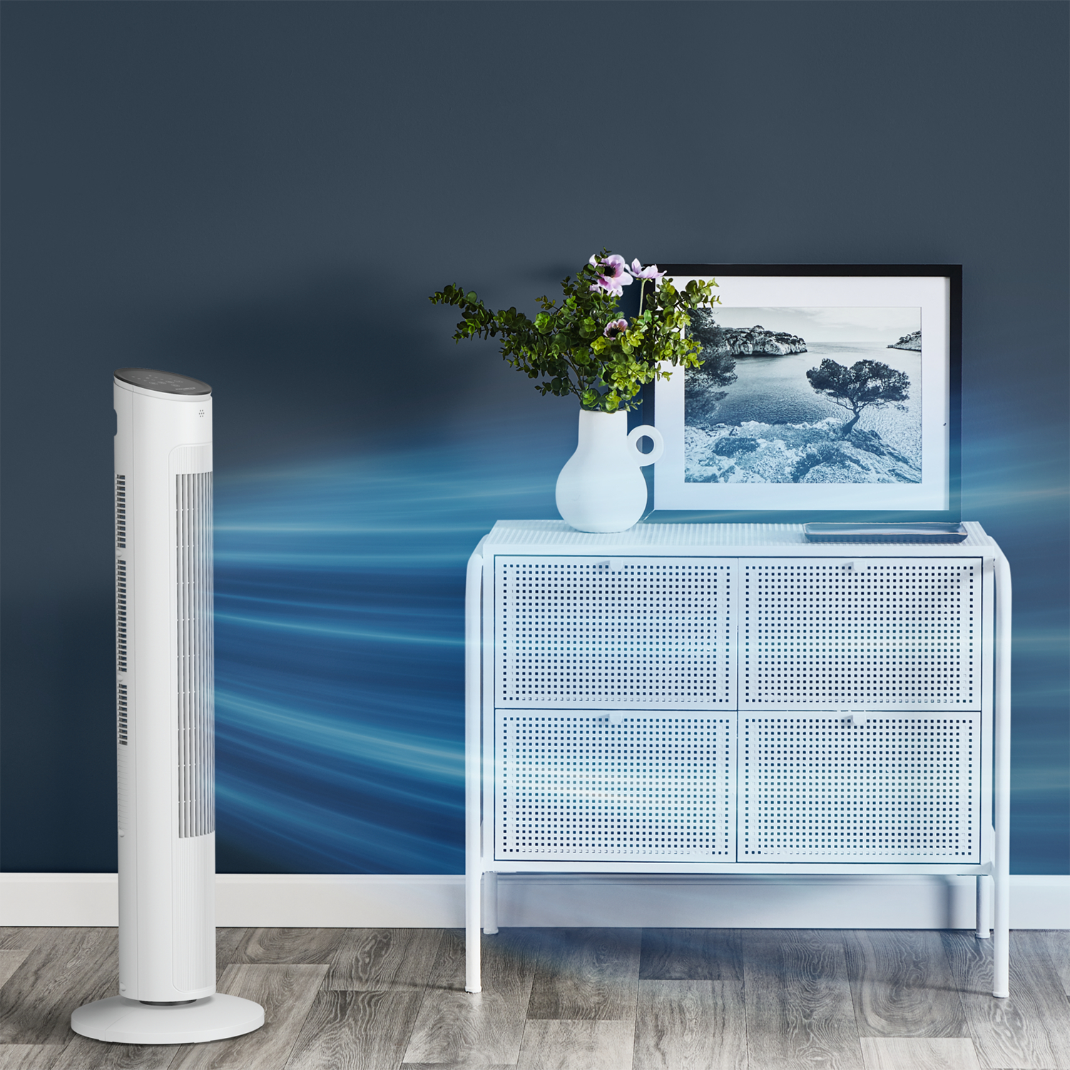 Un ventilateur blanc est à côté d'une armoire blanche avec un cadre photo, un vase et des fleurs. Le fond est un mur bleu.
