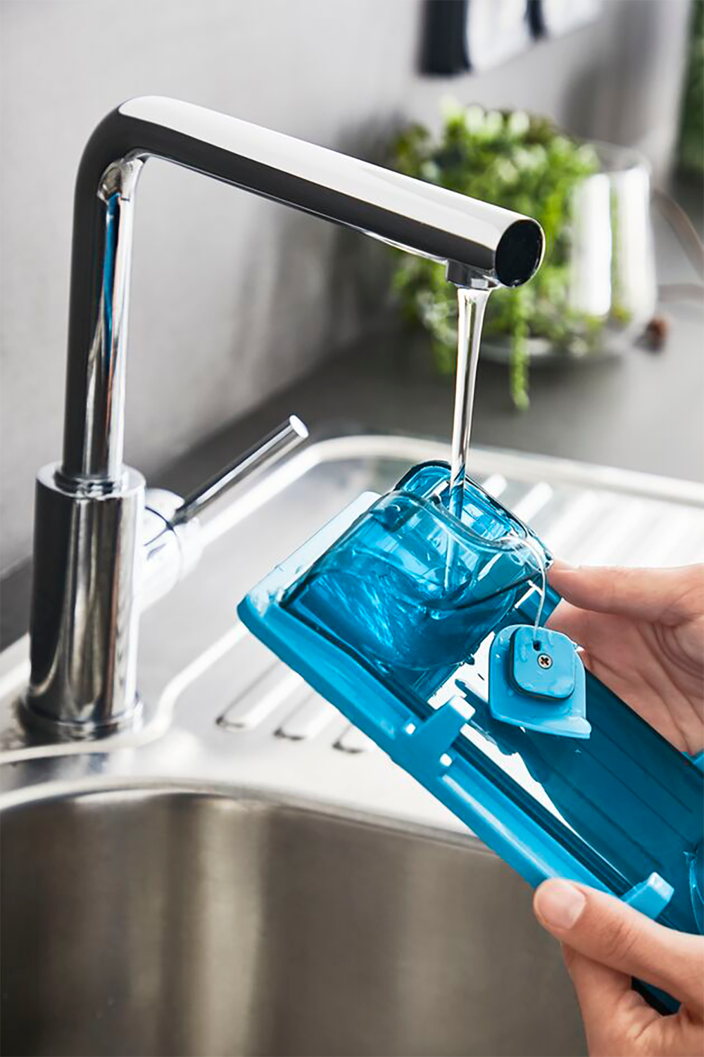 A blue vegetable cutter is being rinsed under a chrome faucet. A hand holds the cutter.