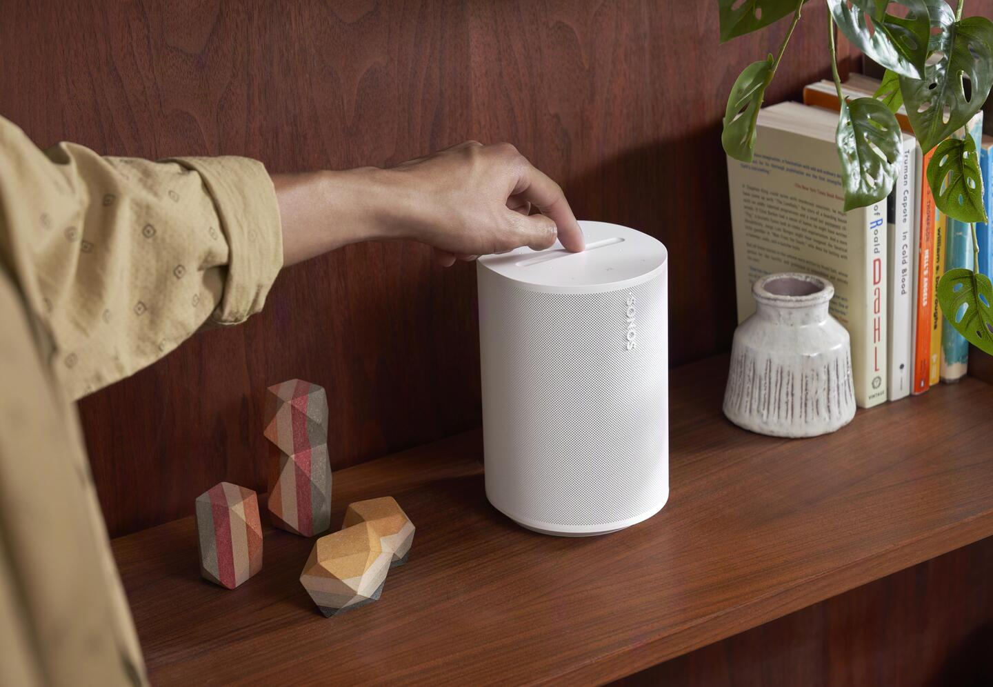 Mano tocando un altavoz Sonos blanco en estante de madera junto a libros.
