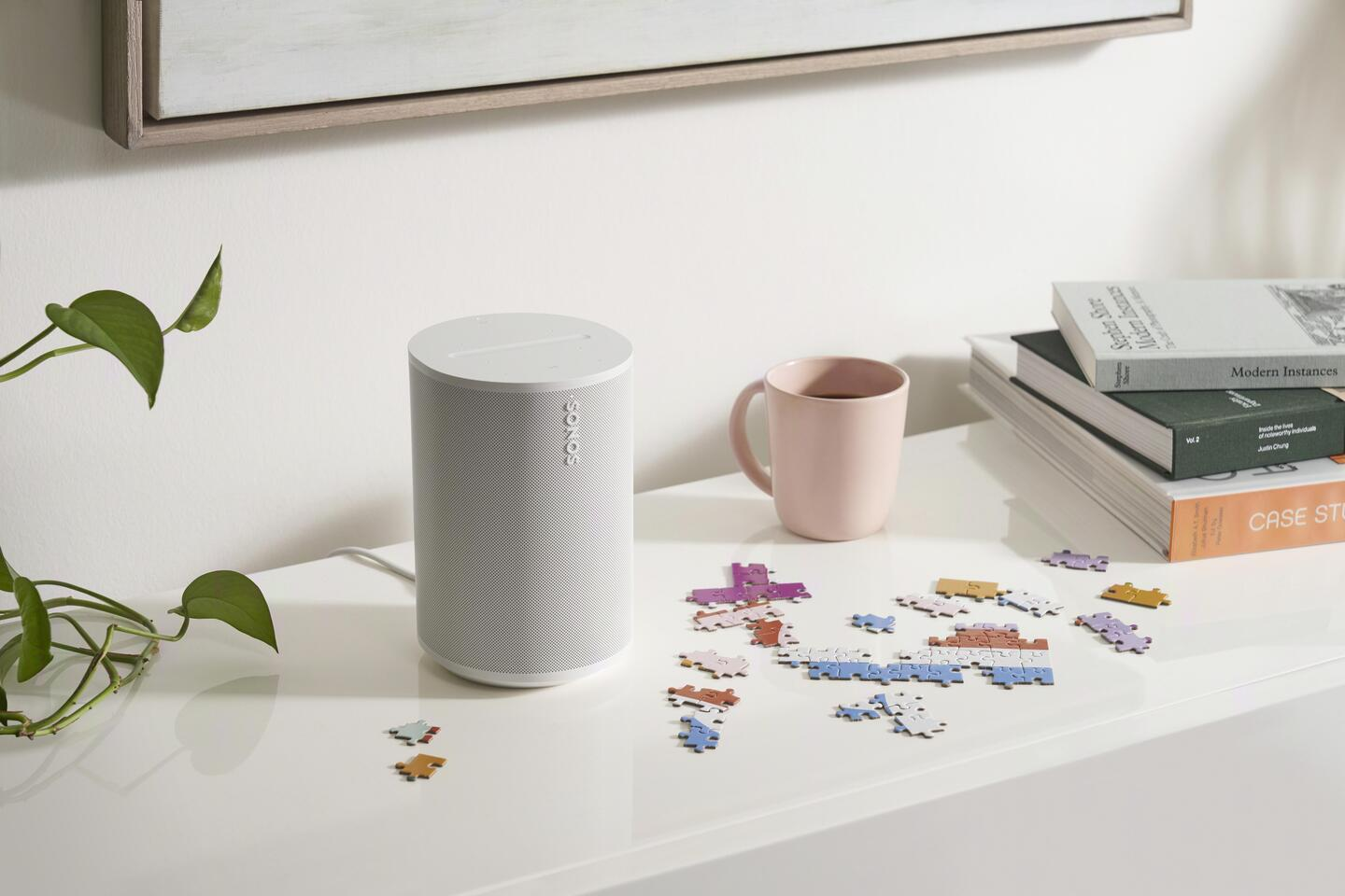 Un altavoz blanco, una taza, libros y un rompecabezas sobre una mesa blanca.
