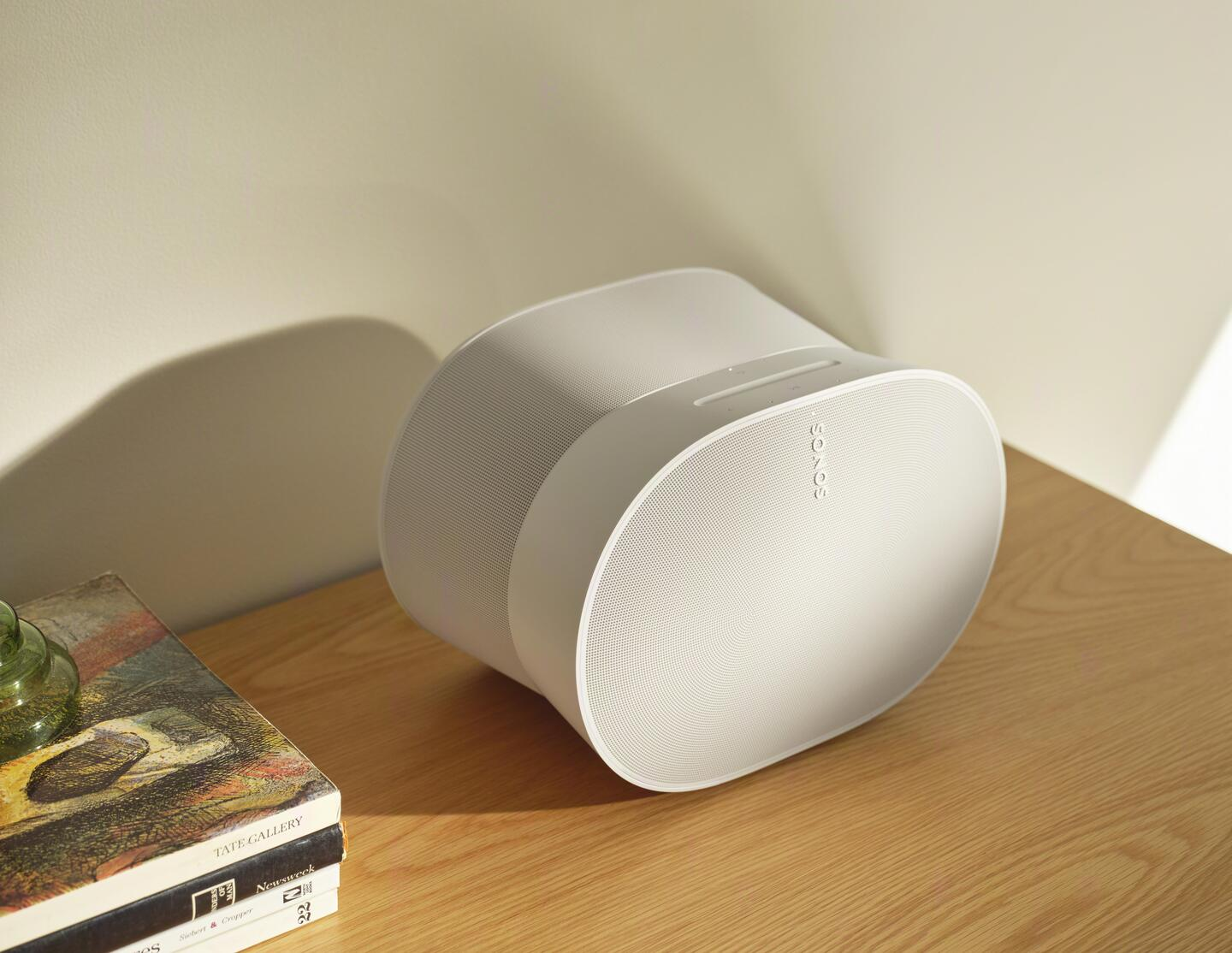 Un altavoz Sonos blanco sobre una superficie de madera. Hay un libro y una pared en el fondo.