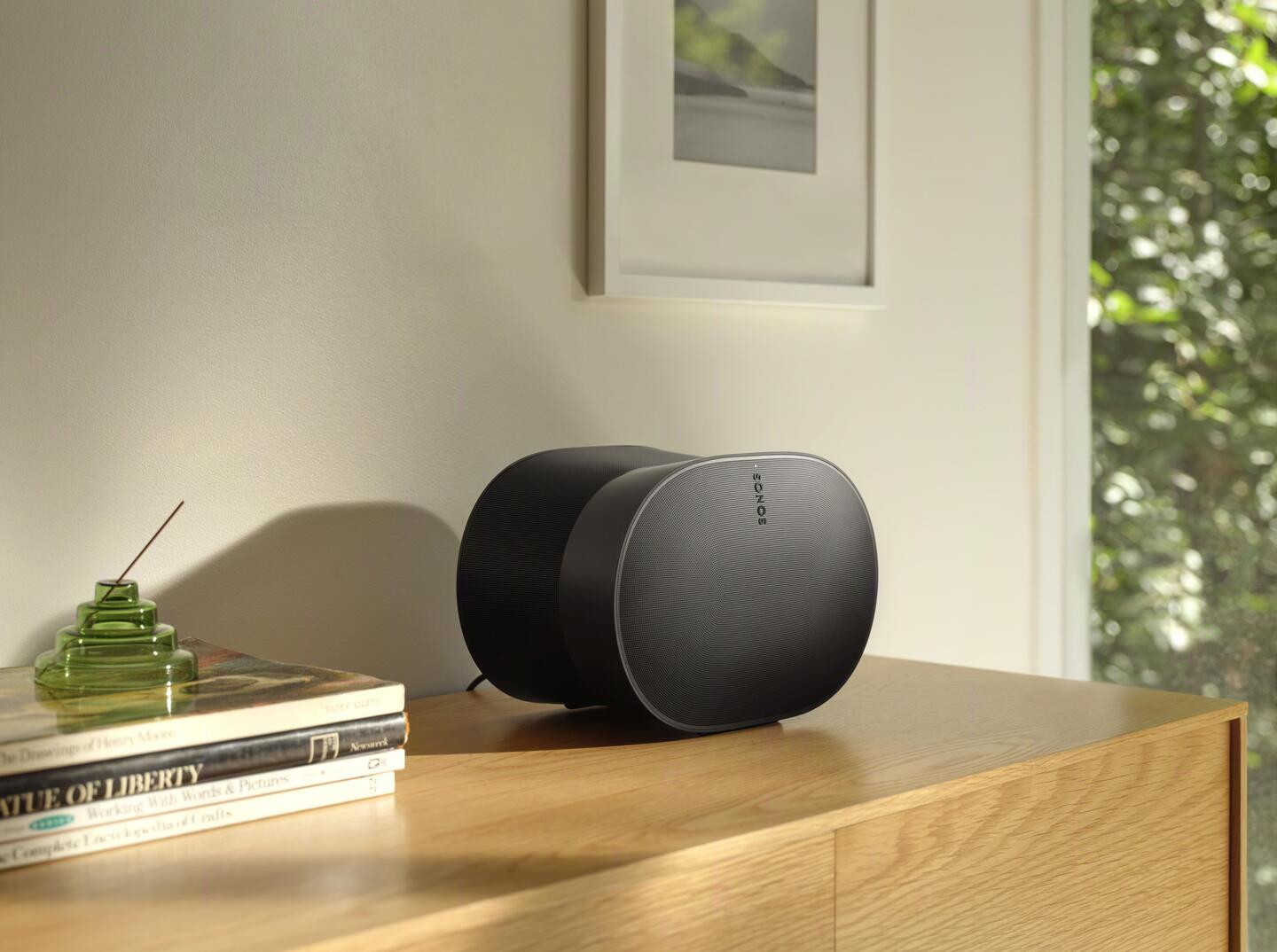 Un altavoz Sonos negro sobre una superficie de madera con libros y una imagen.