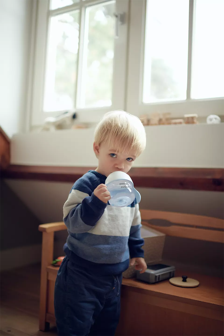 Egy fiatal, szőke hajú fiú kék itatópohárból iszik, egy ablak és egy fa pad közelében állva.
