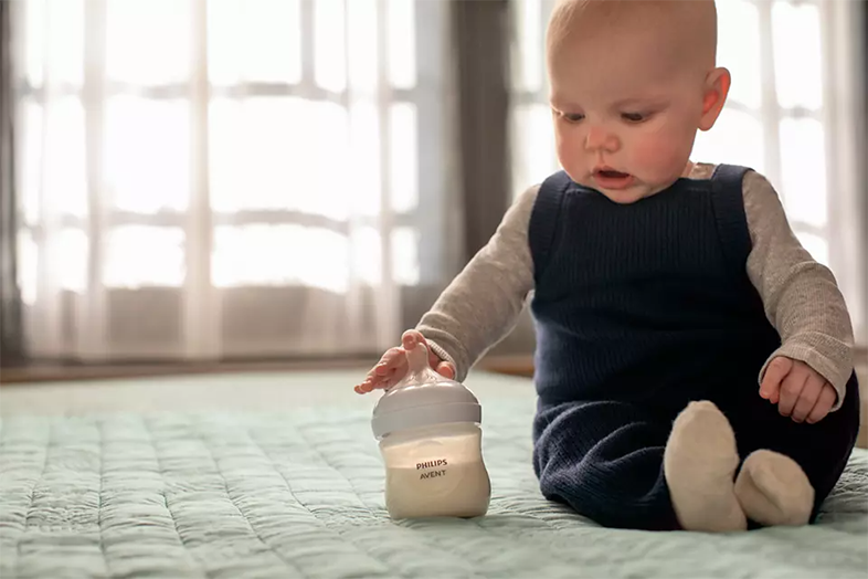 Egy baba a földön ülve egy Philips Avent cumisüveget tart.