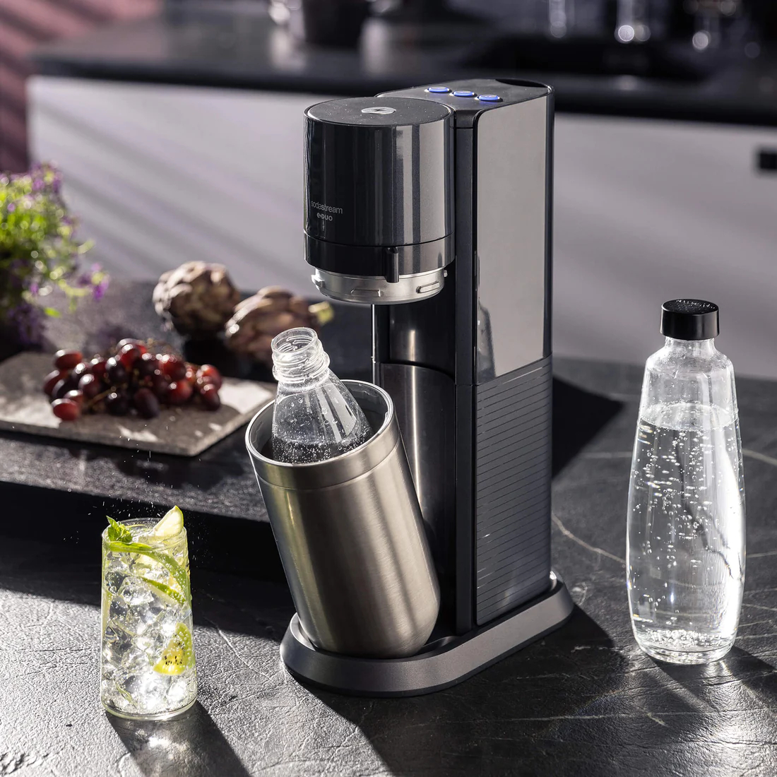 Soda-Maker mit einem Glas Getränk. Es steht auf einer schwarzen Oberfläche mit einer Flasche und Obst in der Nähe.