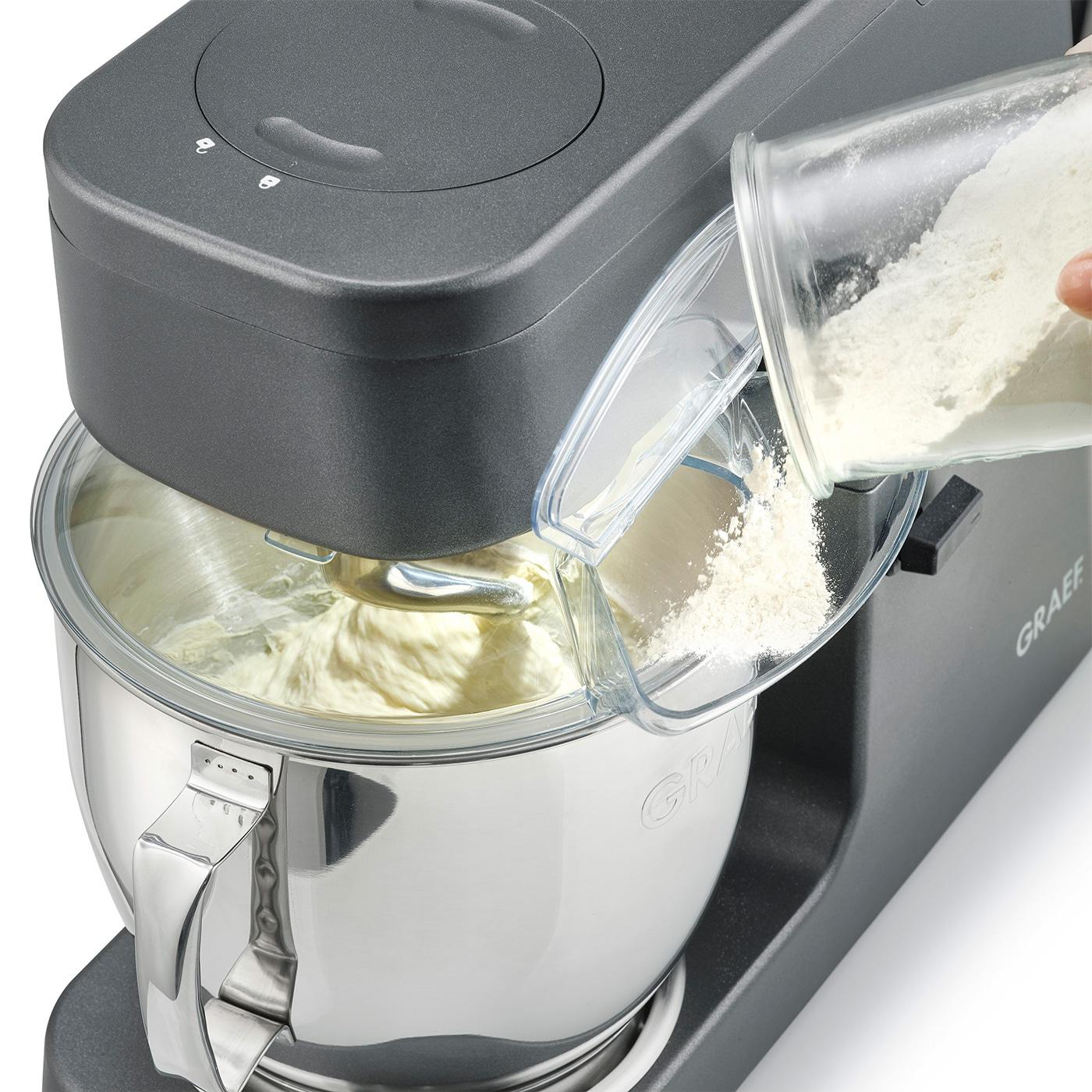 Ein Standmixer mit Teig in der Schüssel, Mehl wird hineingegossen.
