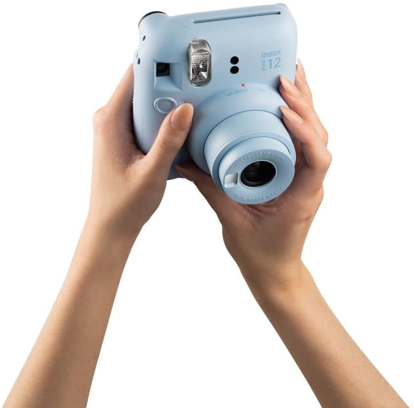 Manos sosteniendo una cámara instantánea Instax Mini 12 celeste, lente hacia adelante.