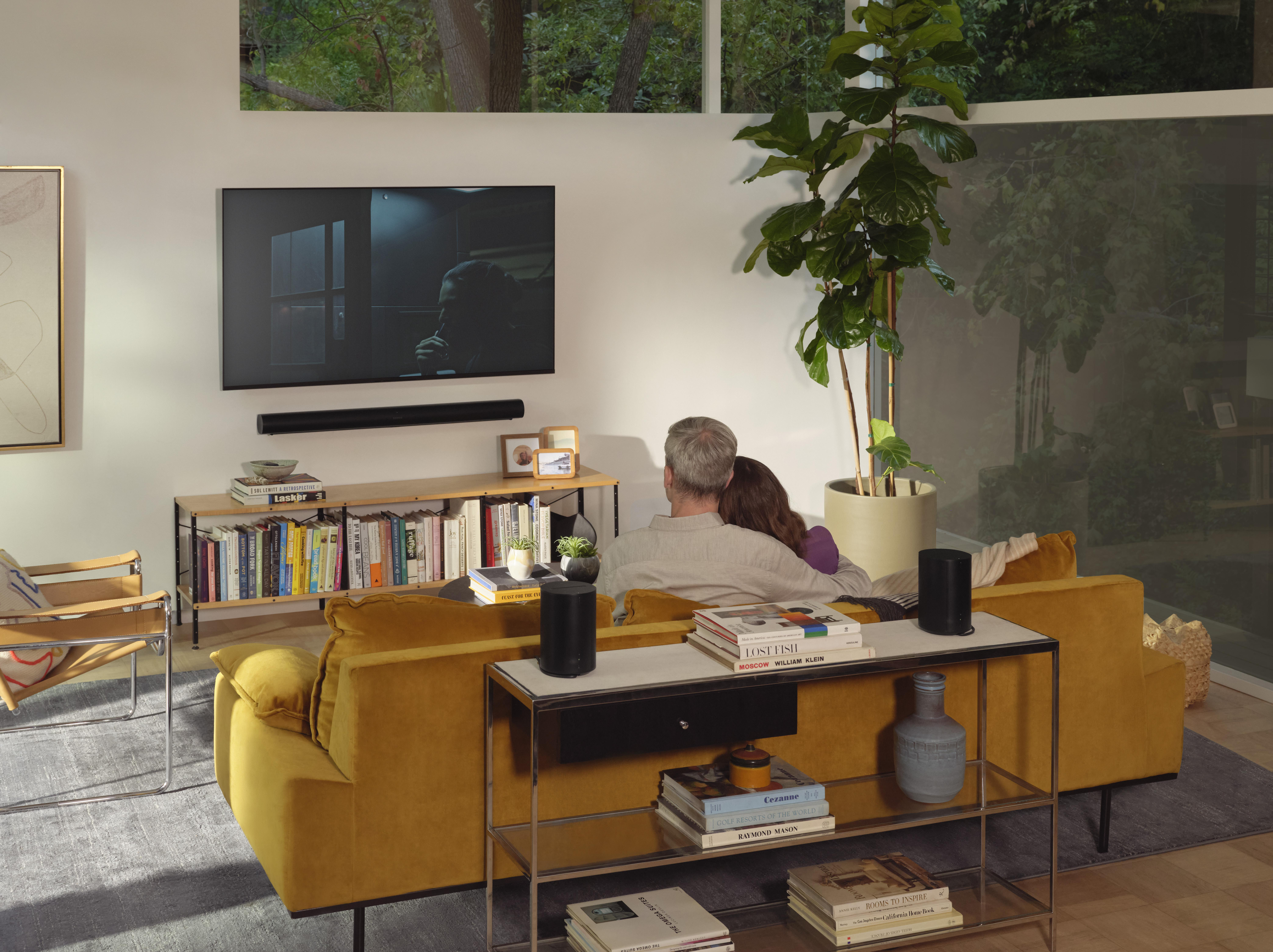 Paar sitzt auf gelbem Sofa und schaut fern. Bücherregal, Lautsprecher und Pflanze sichtbar.