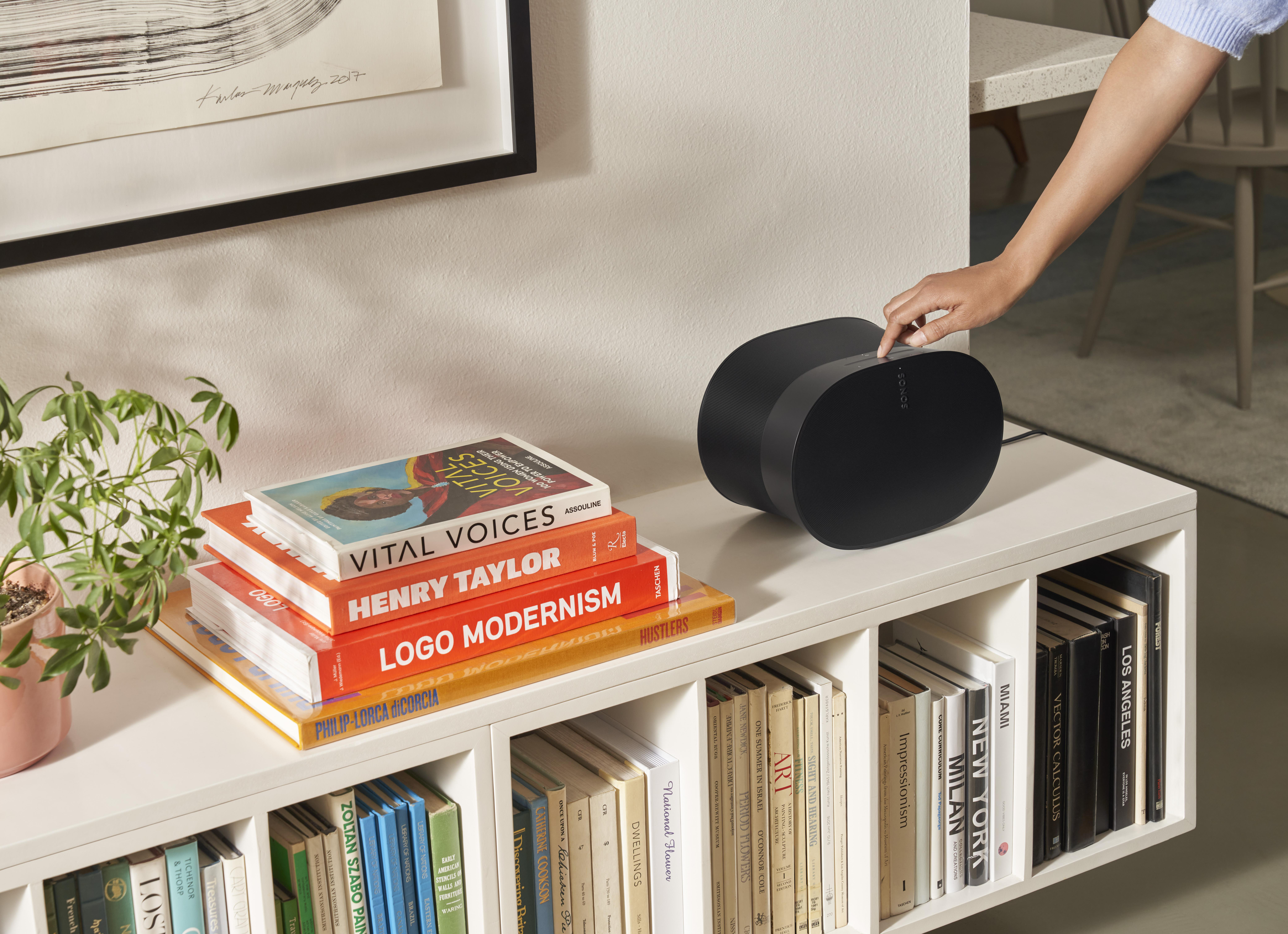 Eine Hand berührt einen schwarzen Lautsprecher auf einem Regal mit Büchern.