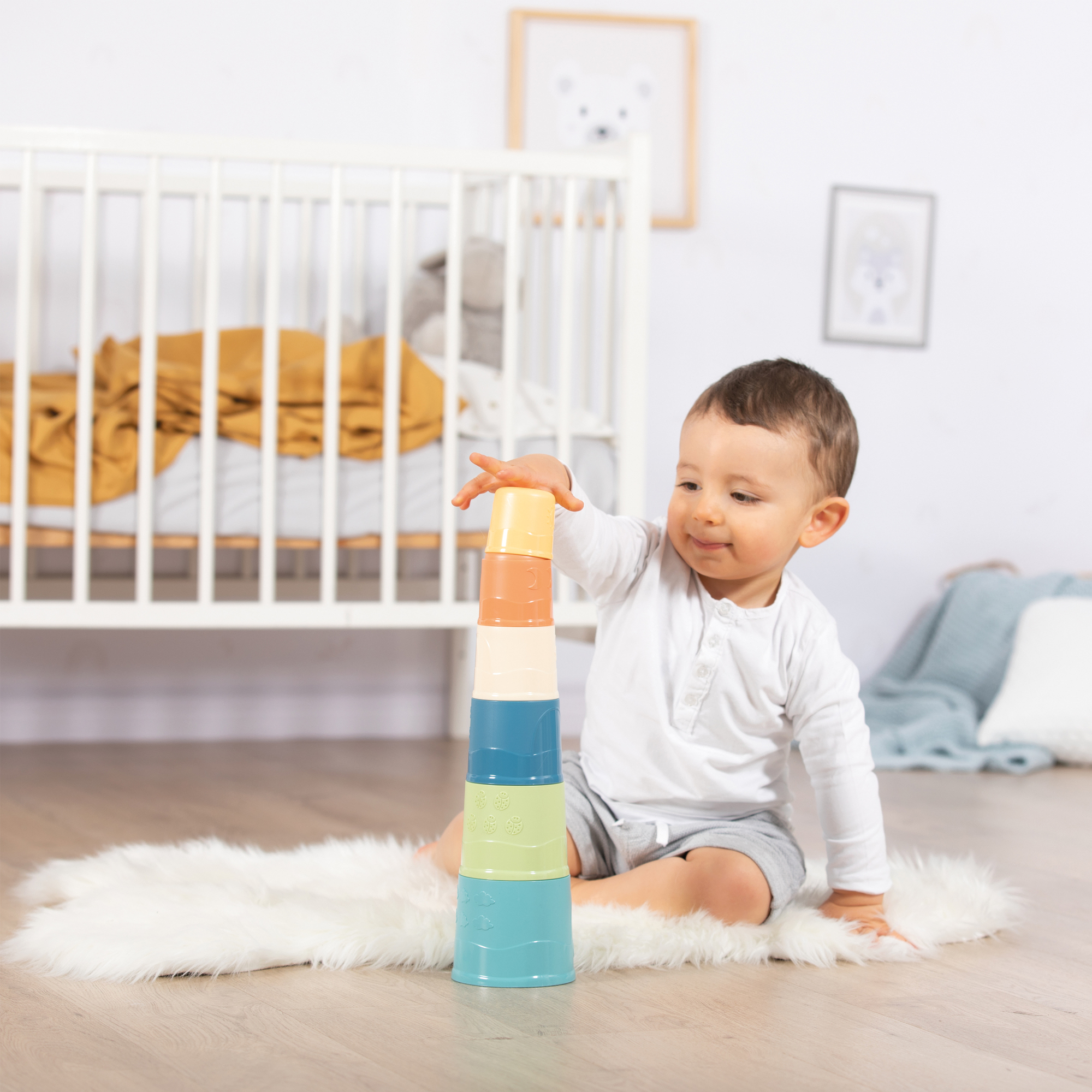 Ein Baby stapelt bunte Becher auf einem flauschigen, weißen Teppich. Ein Kinderbett im Hintergrund.