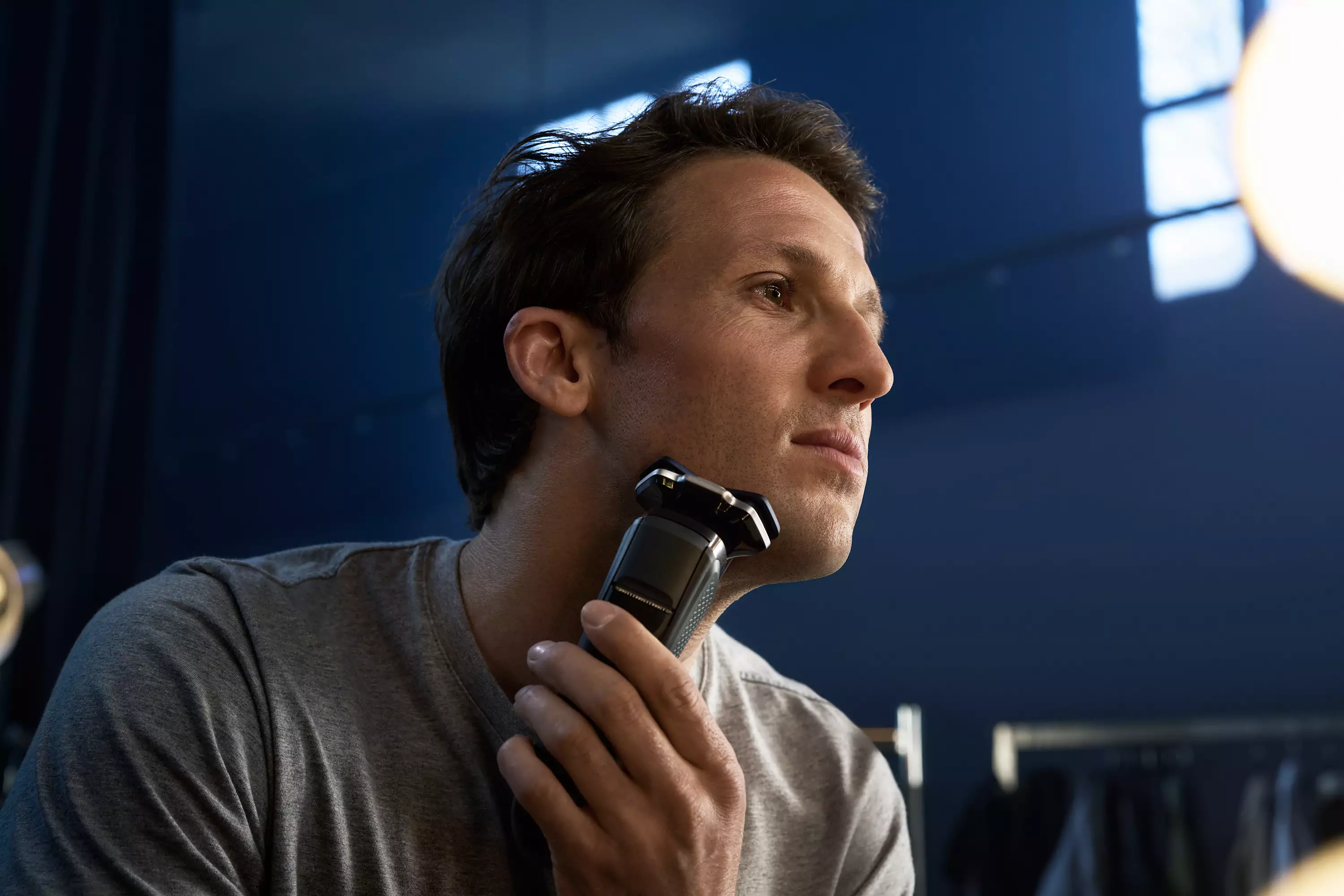 Homme se rase avec rasoir électrique noir. Fond bleu foncé, reflet de lumière.