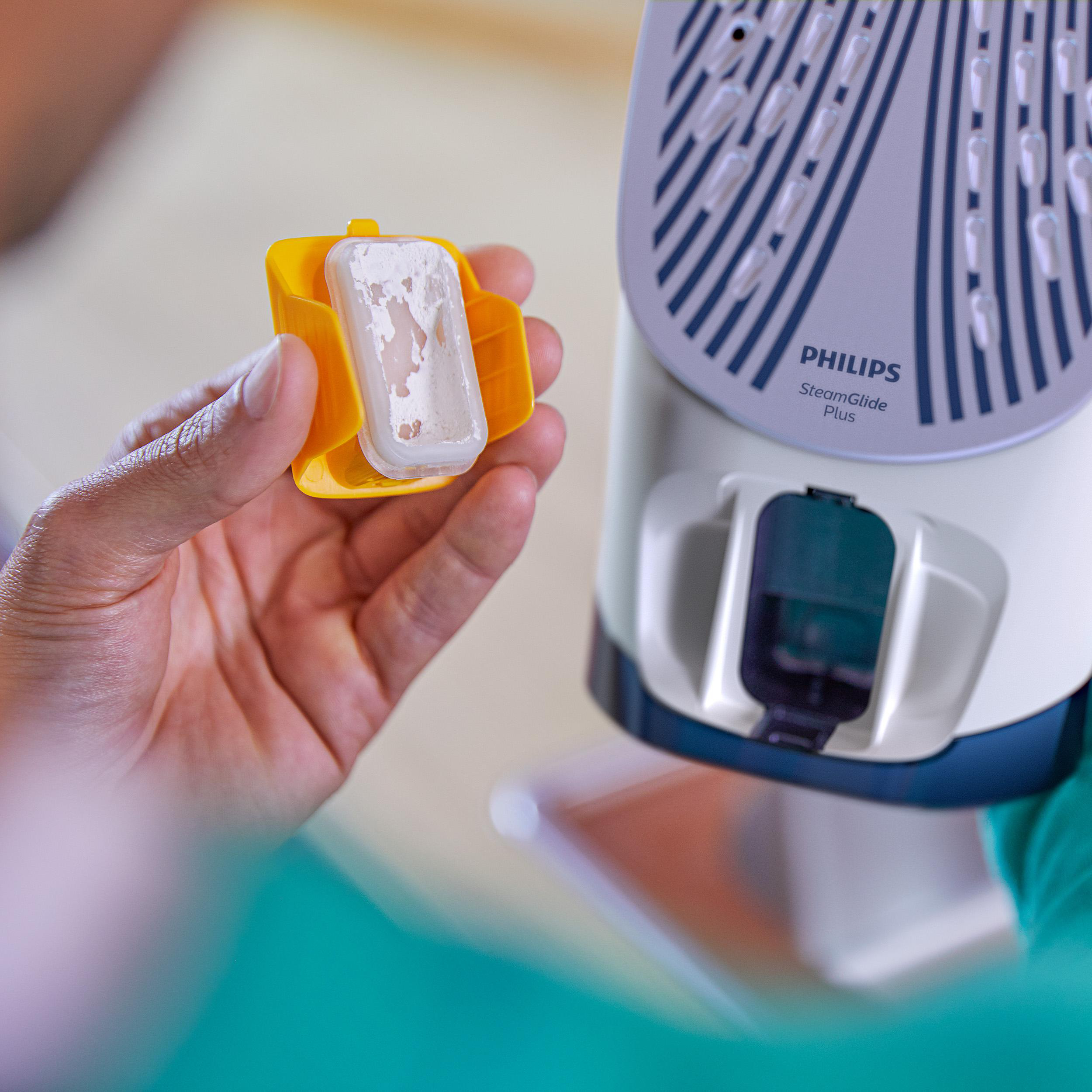 Hand hält gelben Behälter mit weißer, gebrauchter Entkalkungskartusche, neben Philips Bügeleisen.
