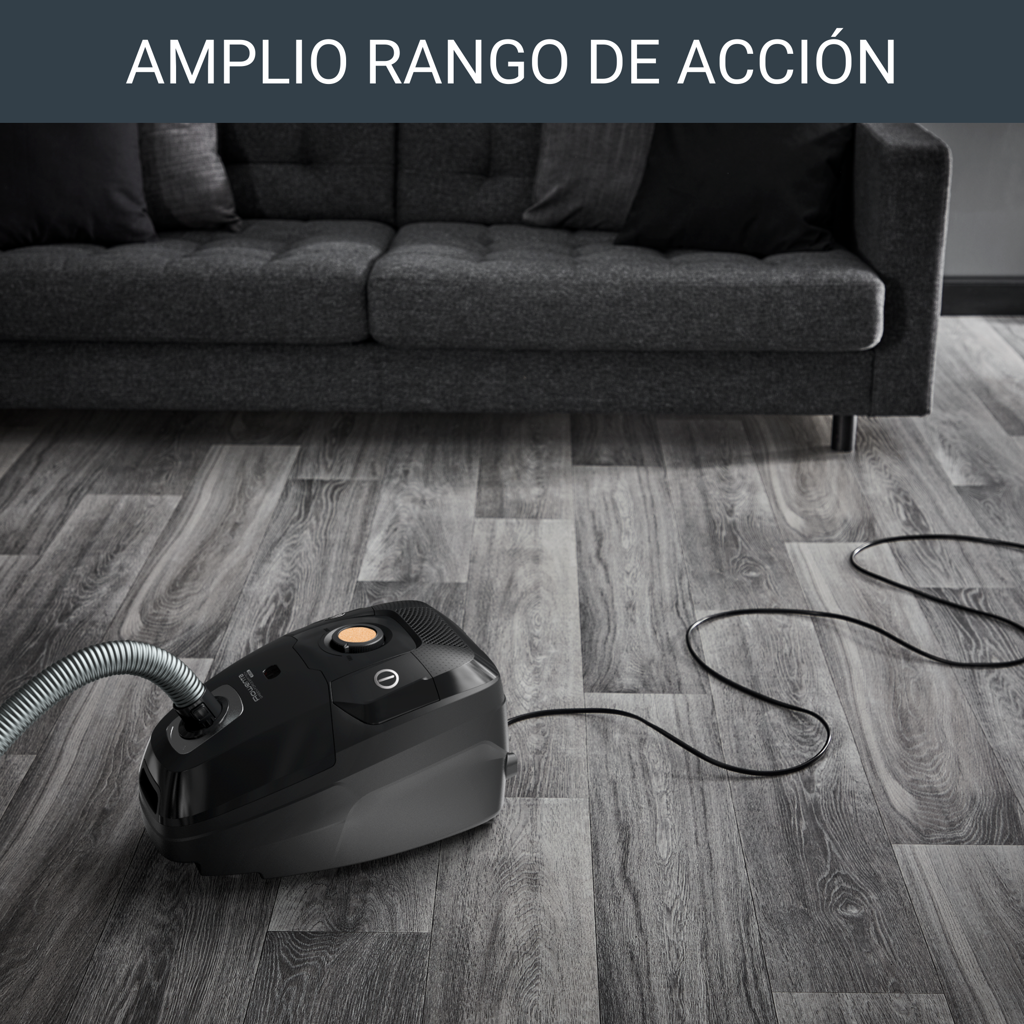 Aspiradora negra en un piso de madera. Un sofá gris está en el fondo.