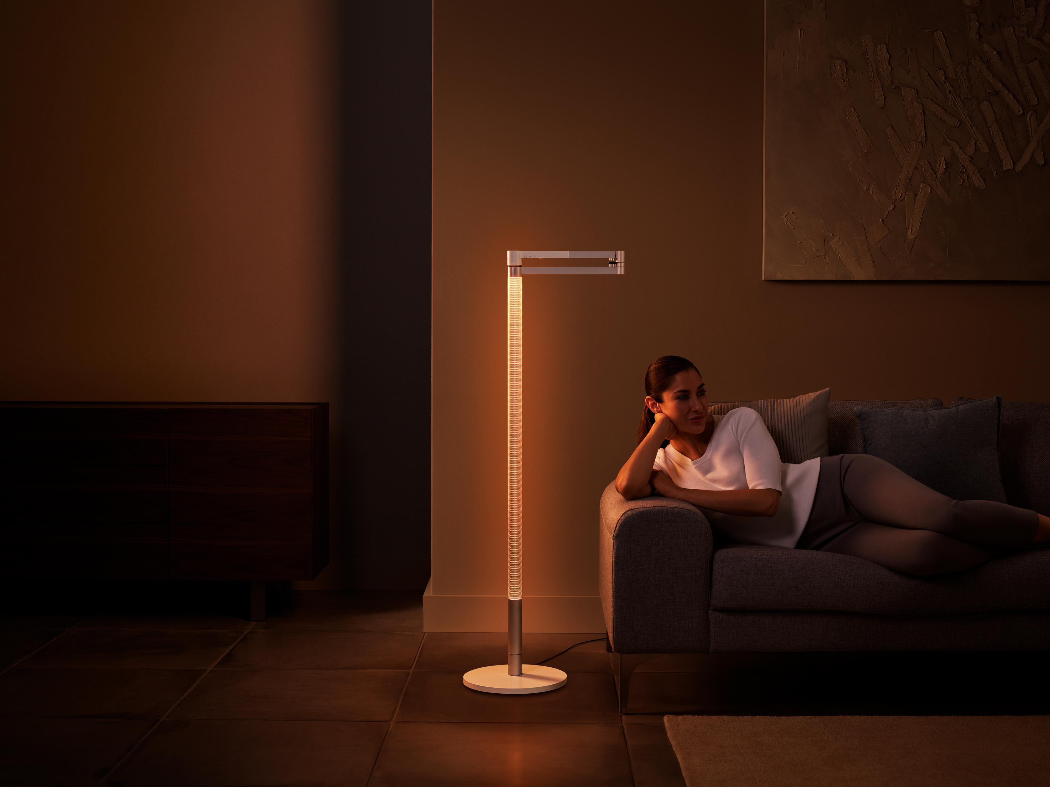 Frau liegt auf einem grauen Sofa, neben einer hohen Lampe. Die Lampe ist an. Der Hintergrund ist braun.